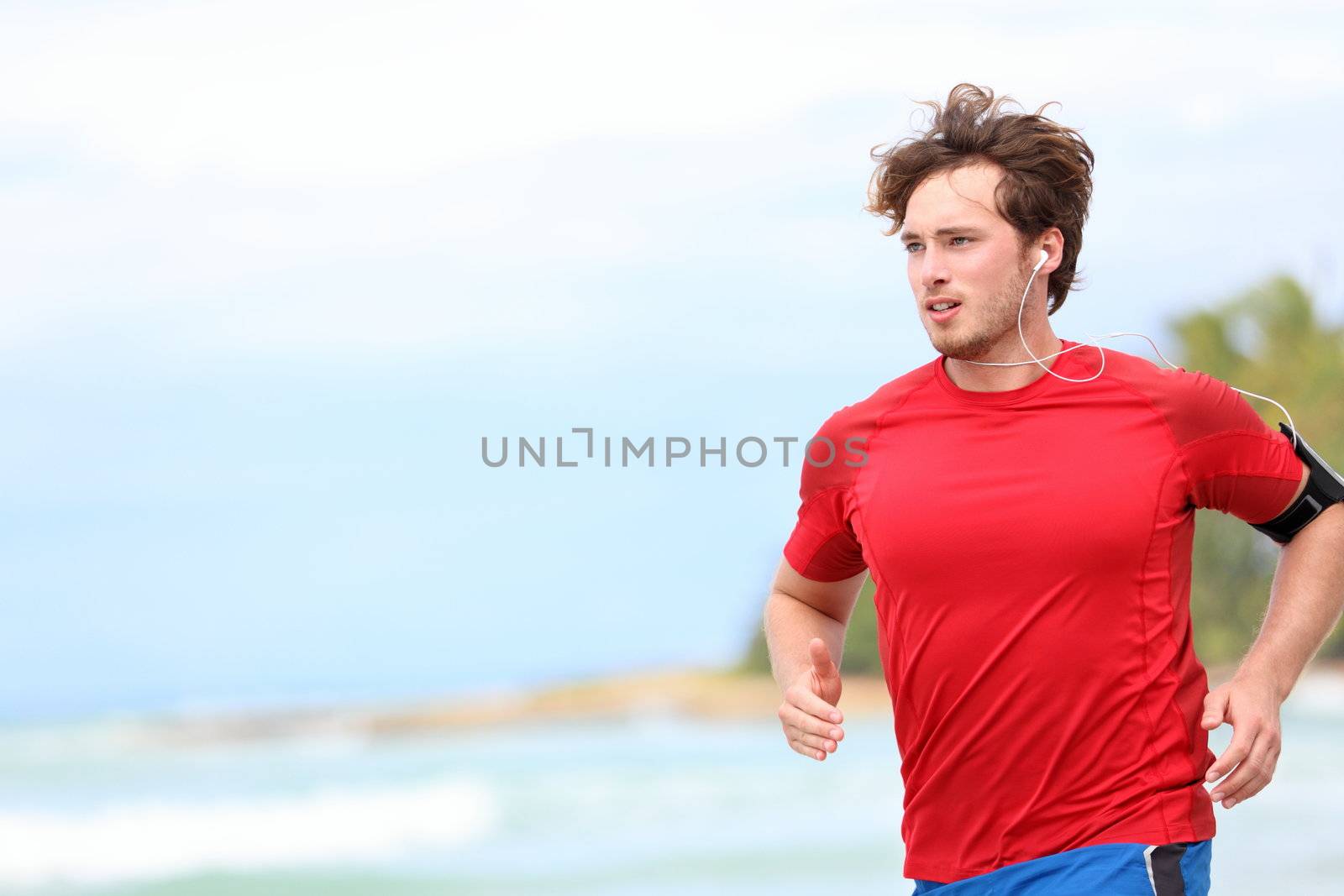 Man jogging on beach. Male runner running listening to music on mp3 player or smart phone. Young caucasian male fitness sport model in red t-shirt.