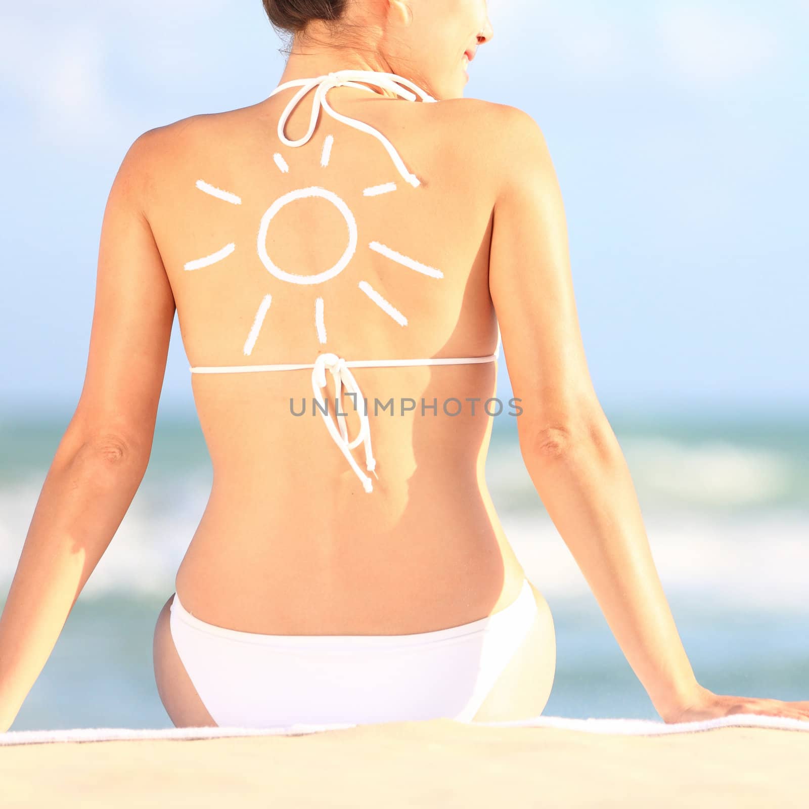 Sunscreen / sun tan lotion sun drawing on woman back. Girl in bikini sitting on beach in sunlight.