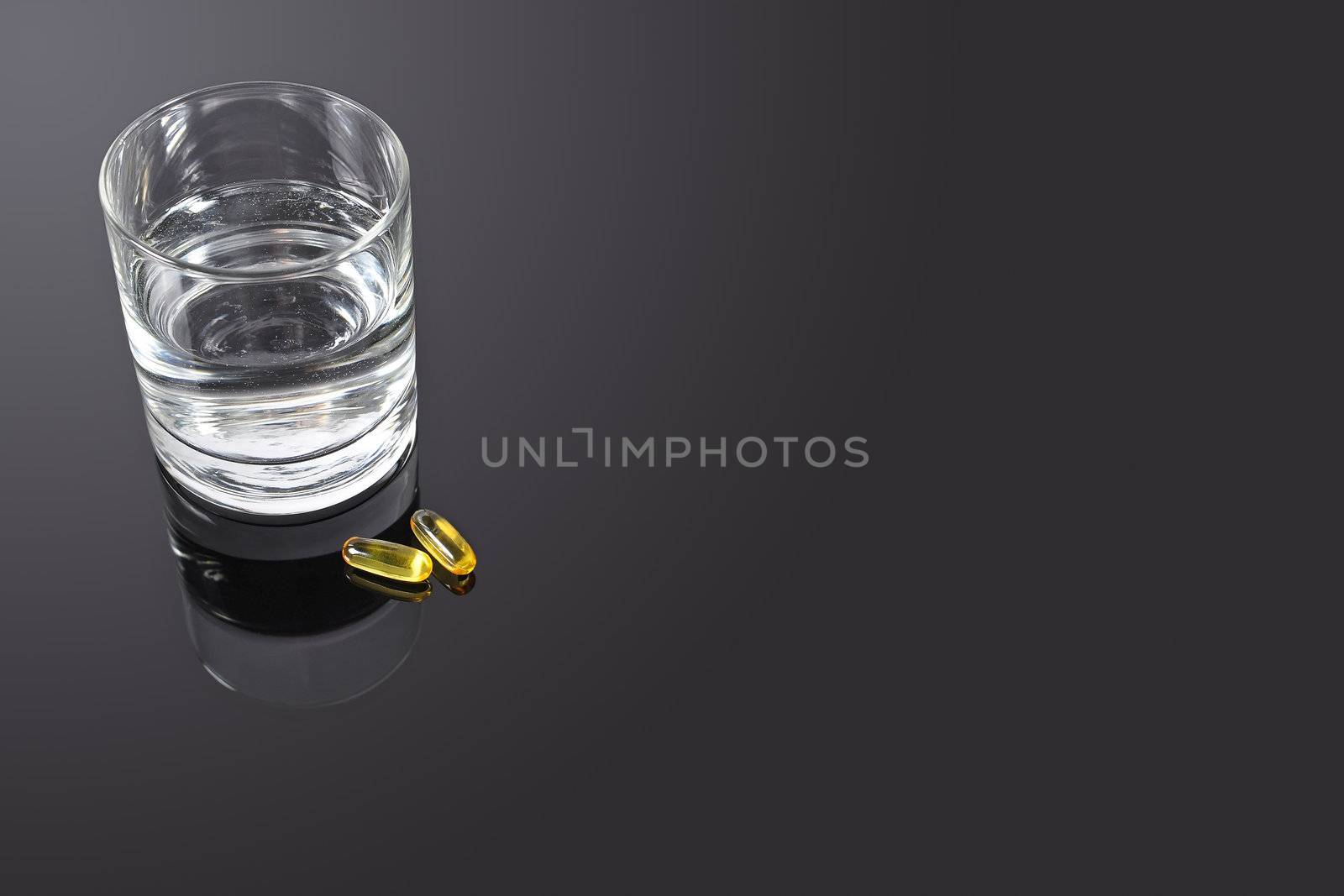 A couple of yellow vitamin pills on a black glass table with a glass of water at the side