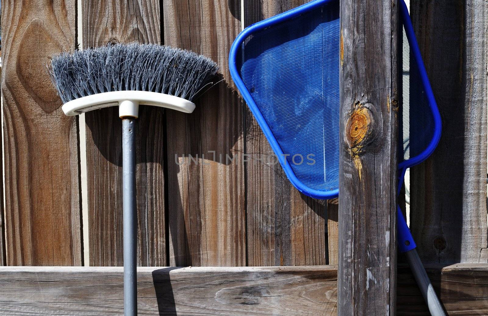 Broom and swimming pool net leaning on a backyard fence.