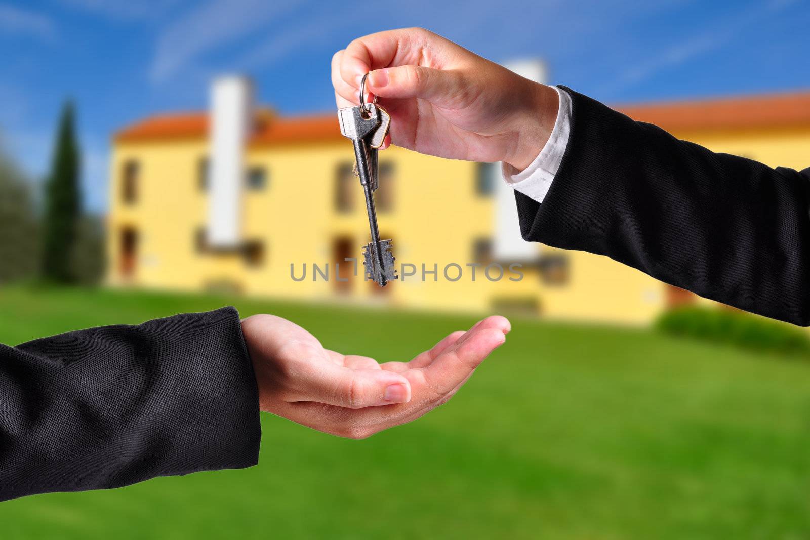 A hand giving a key to another hand. Both persons in suits. House in the background.