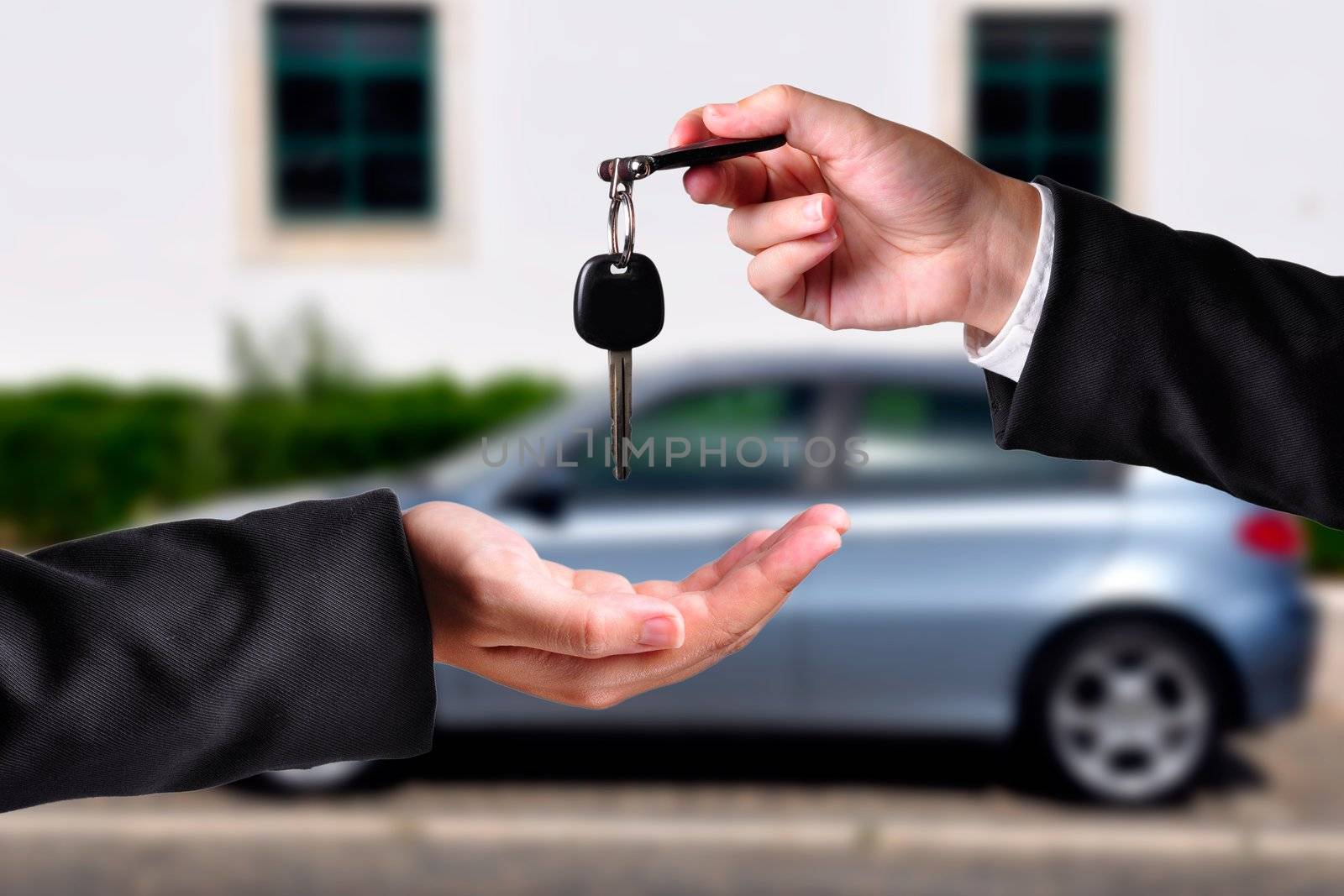 A hand giving a key to another hand. Both persons in suits. Car in the background.