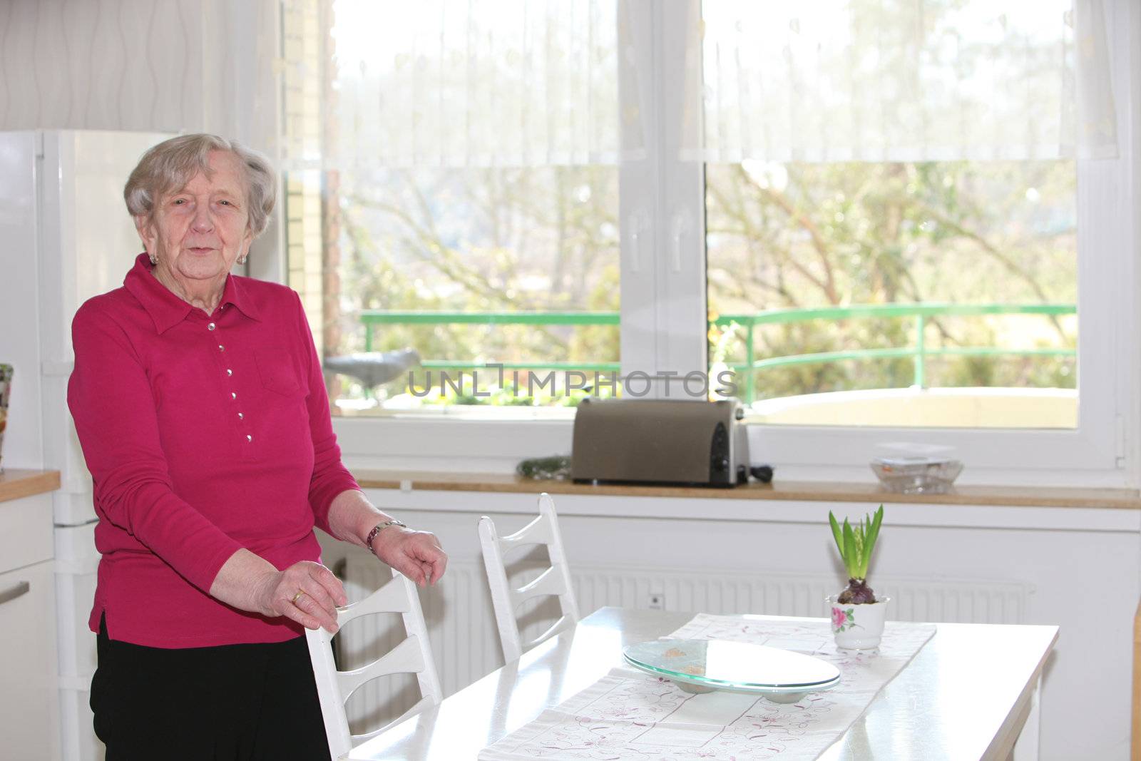 Elderly smiling lady standing next to a white table in front of large light windows