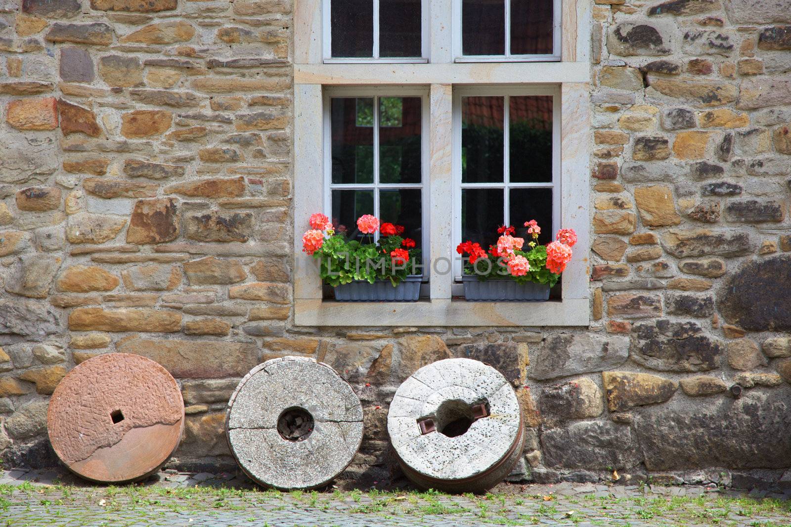 Rustic wooden sash window in an old stone walled building with colourful flower boxes of geraniums and round grindstones balanced on the grass below