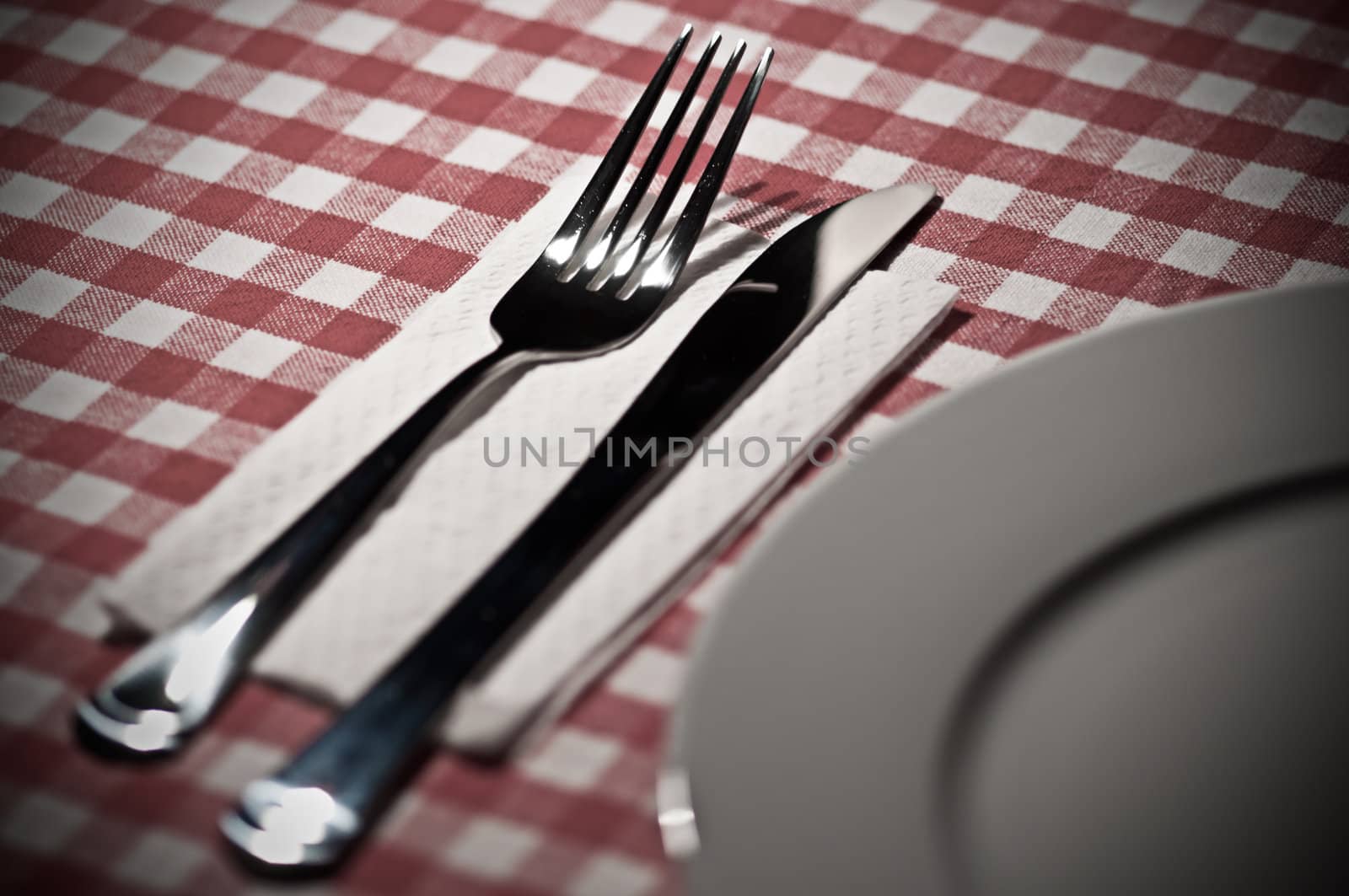 Fork, knife and plate on a table with a red and white tablecloth. Desaturated and gritty look.