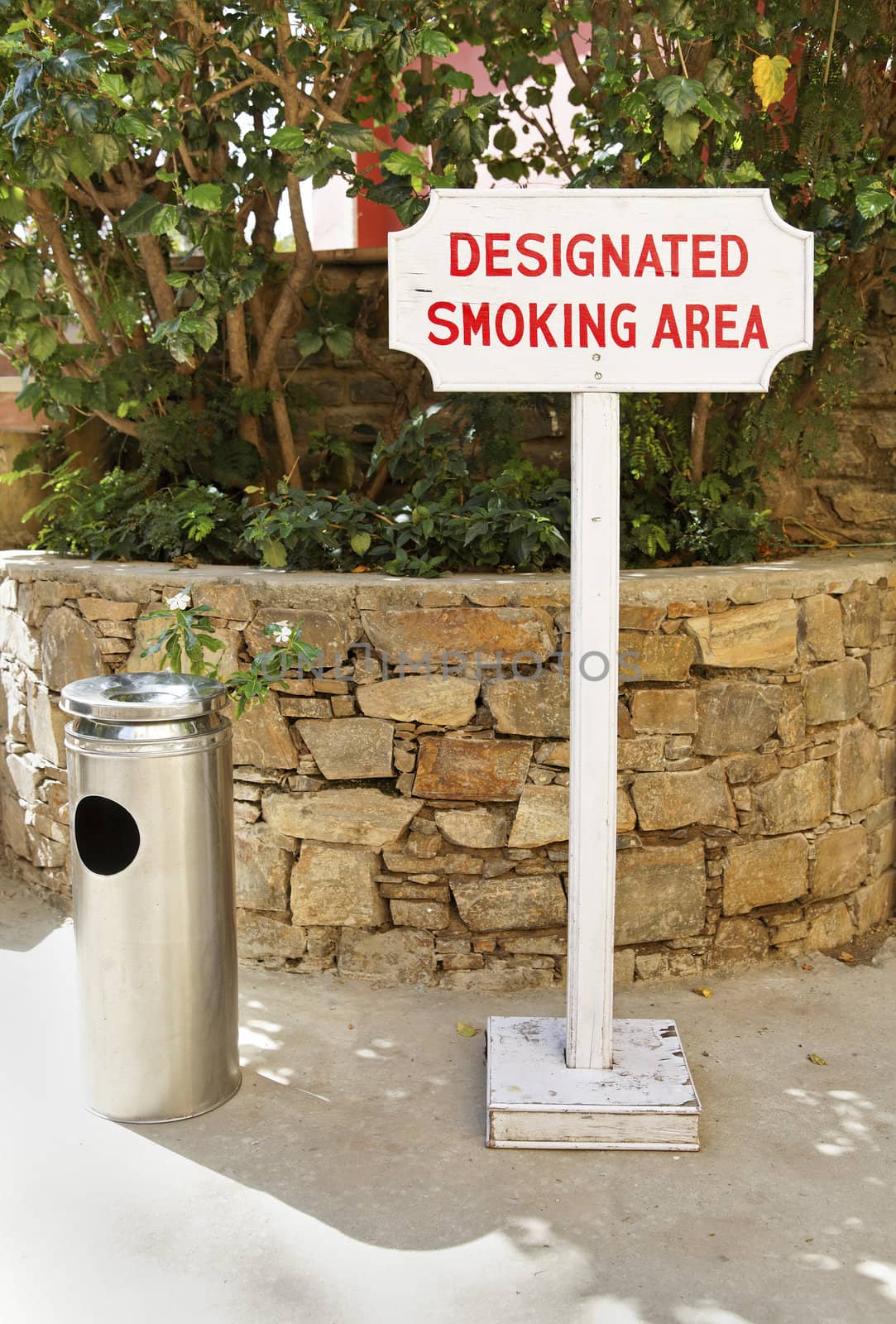 vertical take of a free standing sign and ashtray of a public smoking area in the shade of a garden corner
