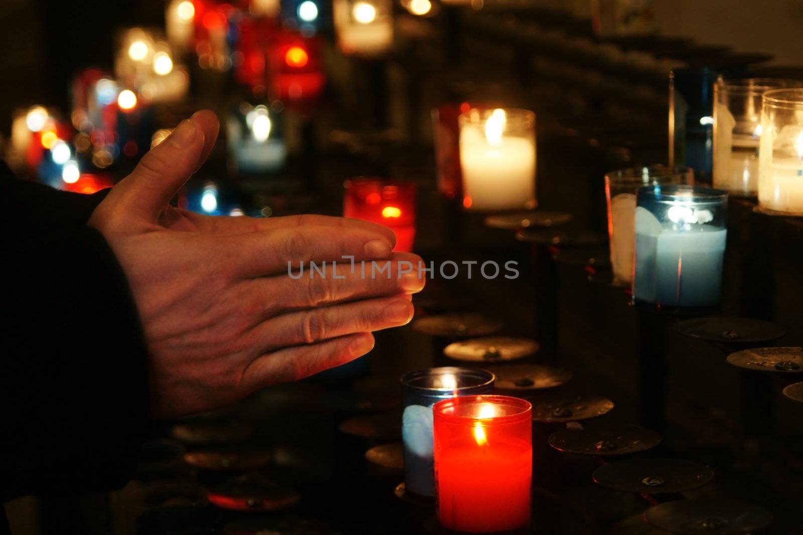 Religion concept: Praying hands in front of a series of colorful glass church candle at a place of worship; soft candlelight lighting.