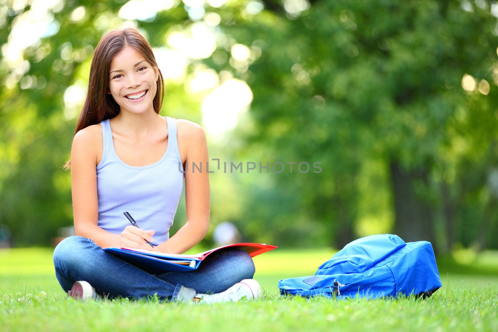 Student studying in park. Joyful happy asian girl student sitting writing and reading outside on university campus or park. Mixed race Chinese Asian / Caucasian female student woman looking at camera.