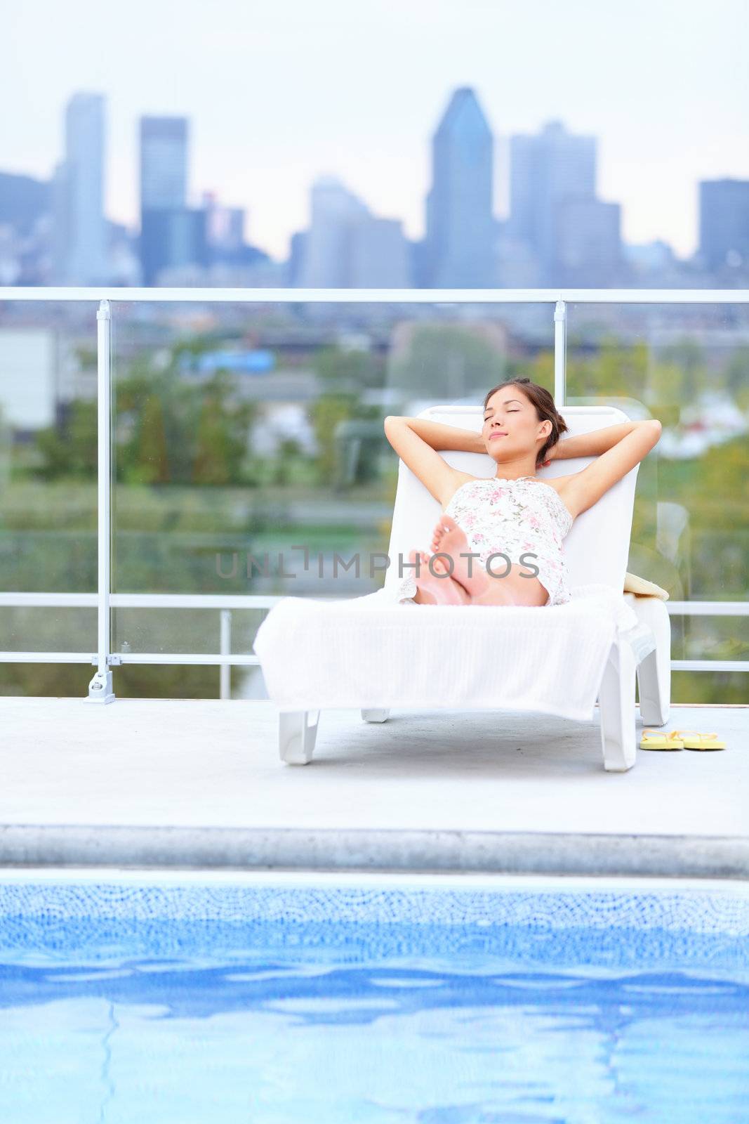 Woman relaxing at rooftop pool in city with skyline in background. Young female model lying down at sun lounger with Montreal skyline in background.