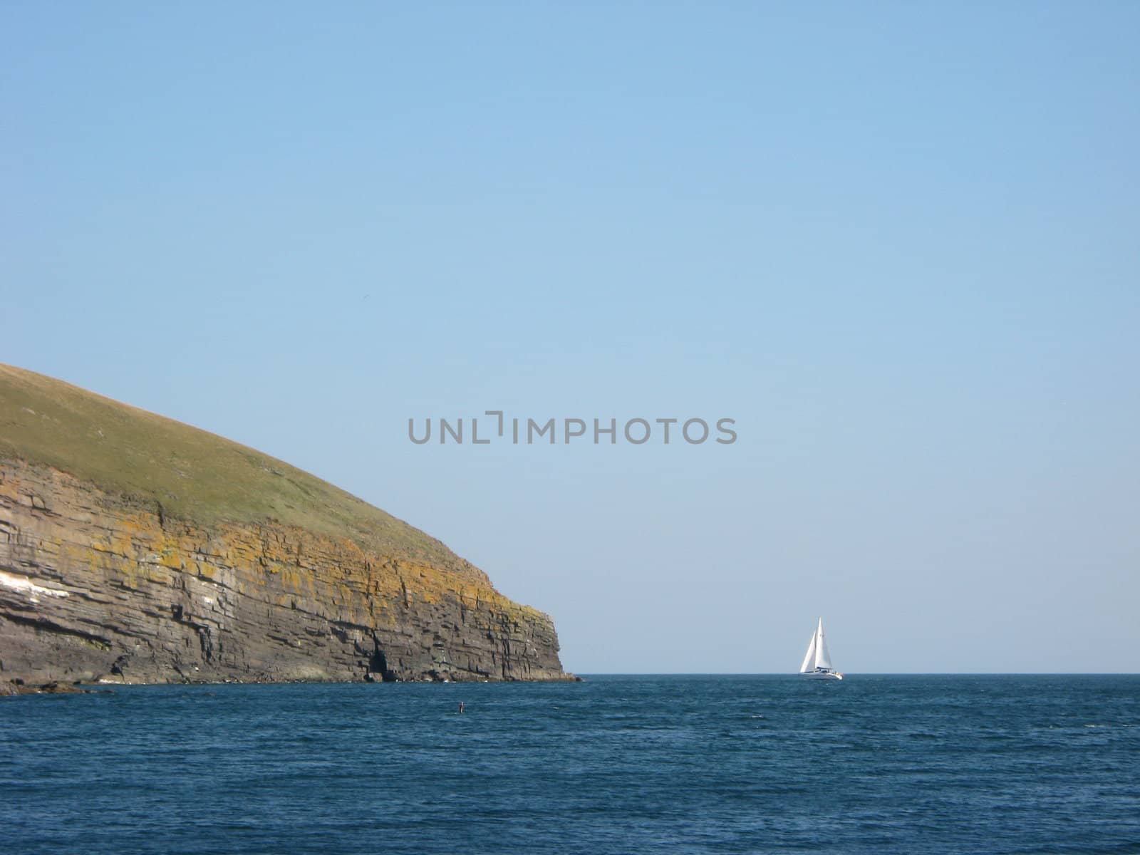 A yacht sailing into the distance around a headland cliff with blue sea and sky.