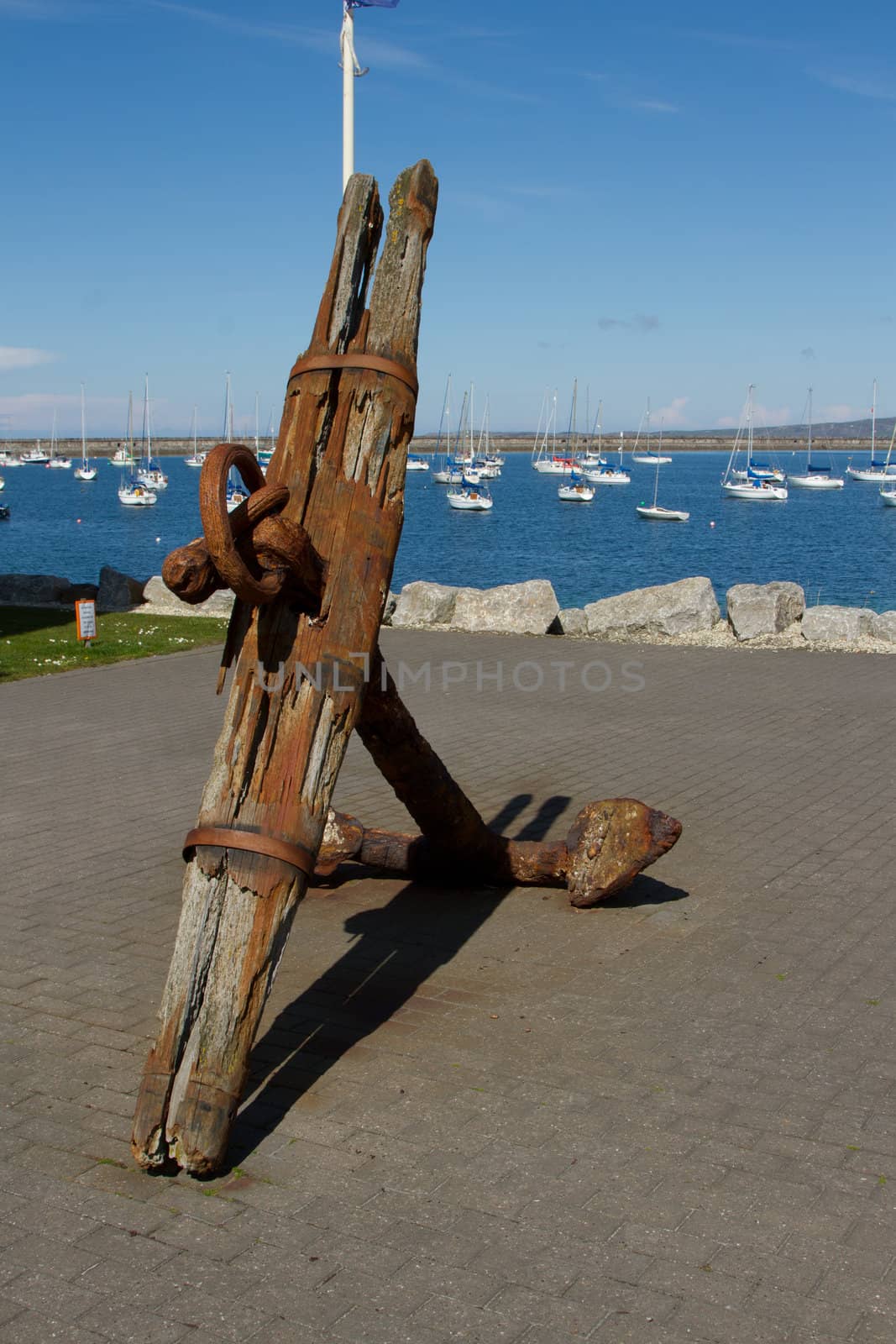 A vintage, antique anchor made of wood and rusty metal on a pavement infront of a marina with yachts.