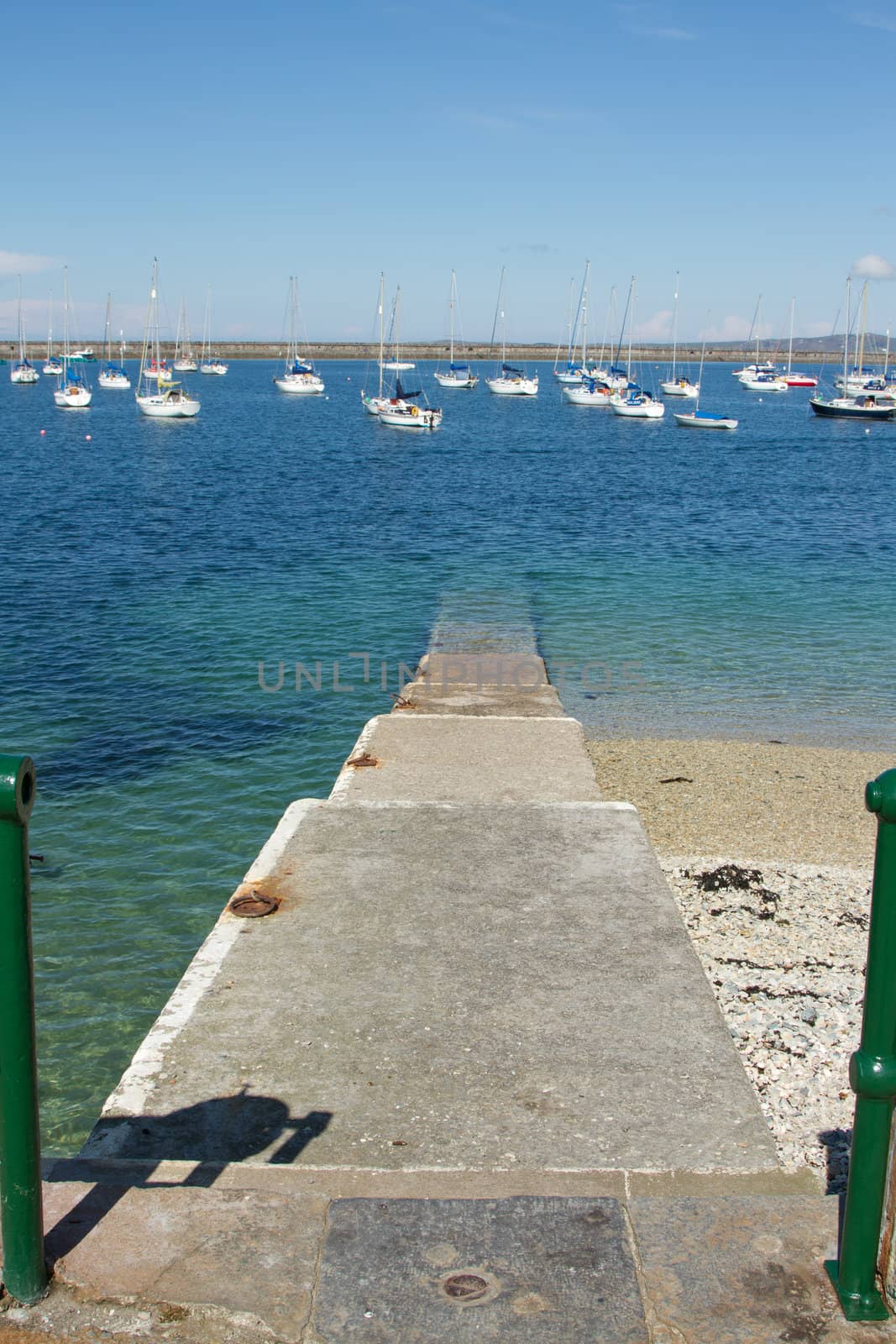 A set of concrete steps leads into the clear waters of a marina with moored yachts and a breakwater in the background.