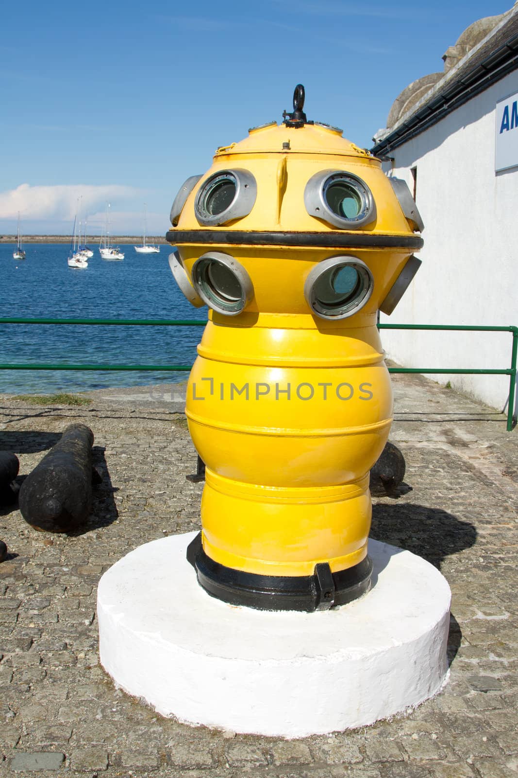 An antique diving bell painted yellow with viewing portals on a white plinth at a quay with the sea in the background.