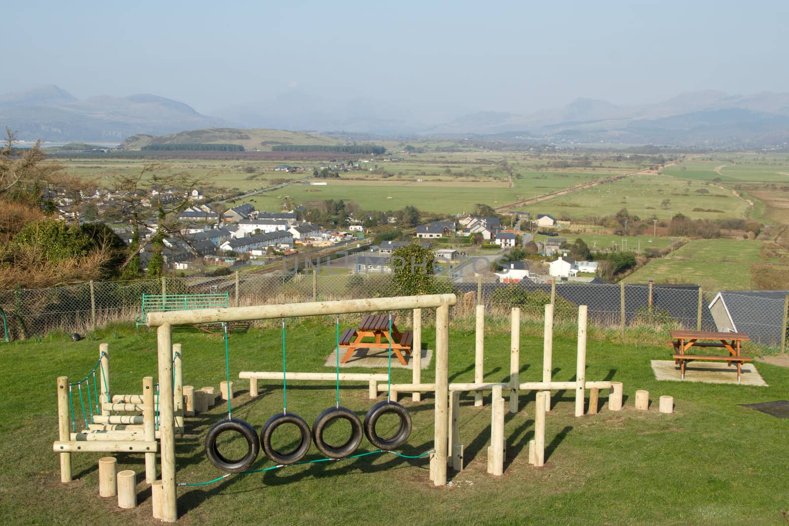 A children's playground with an obstacle course made from wood and tyres on grass in a fenced area with a town and mountains in the distance.