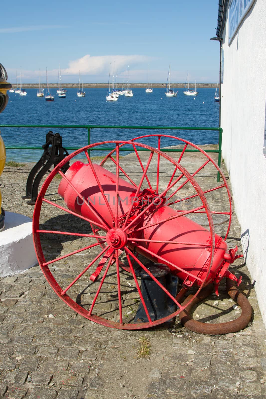 A vintage subaqua air compressor painted red with a tank, wheels and pipe on a cobbled quayside with a marina in the background.