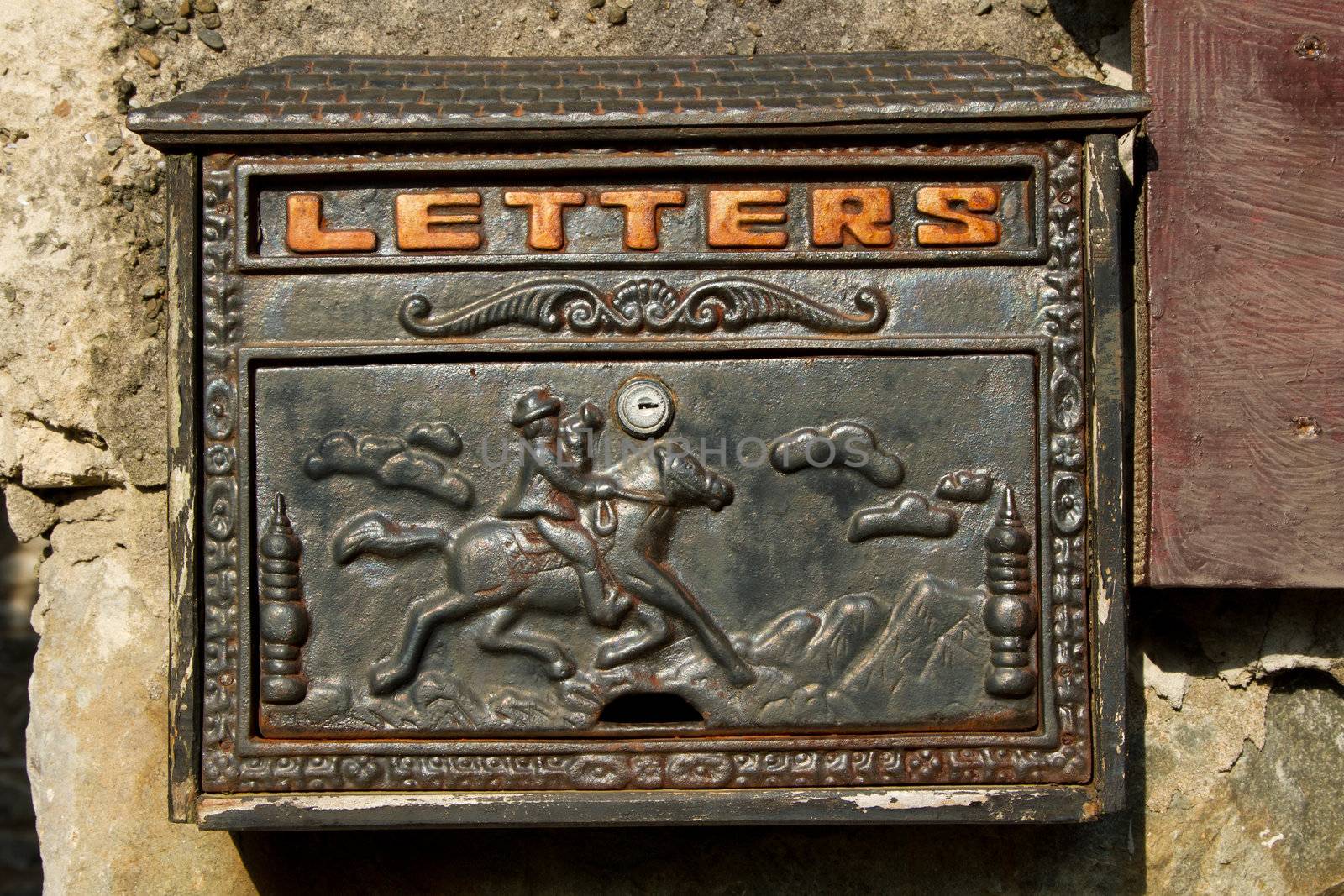 A metal letter box with ornate design painted in black with rust on a stone wall.