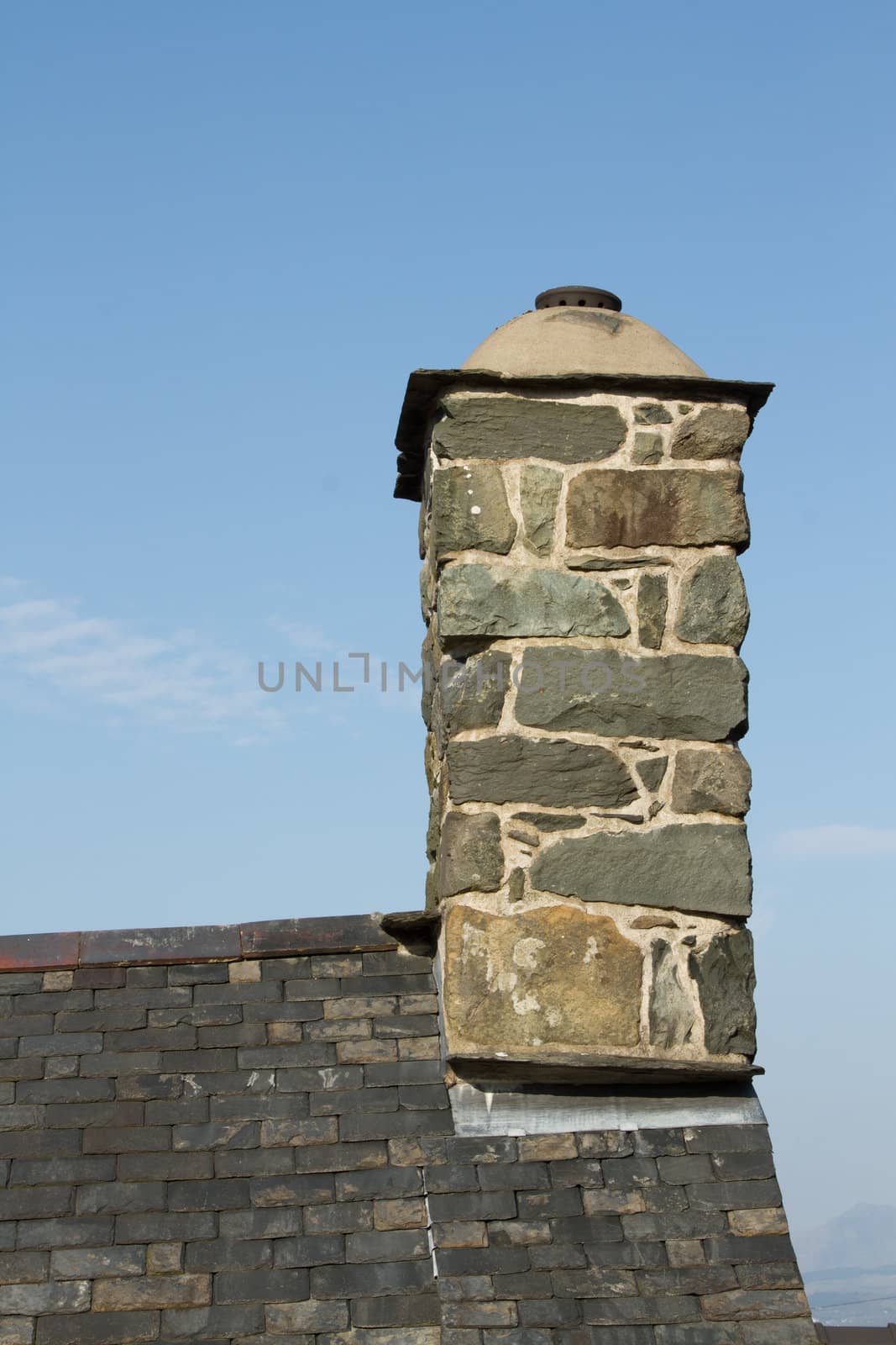 A chimney built from stone with pointing and lead flashing on a slate roof with blue sky in the background.