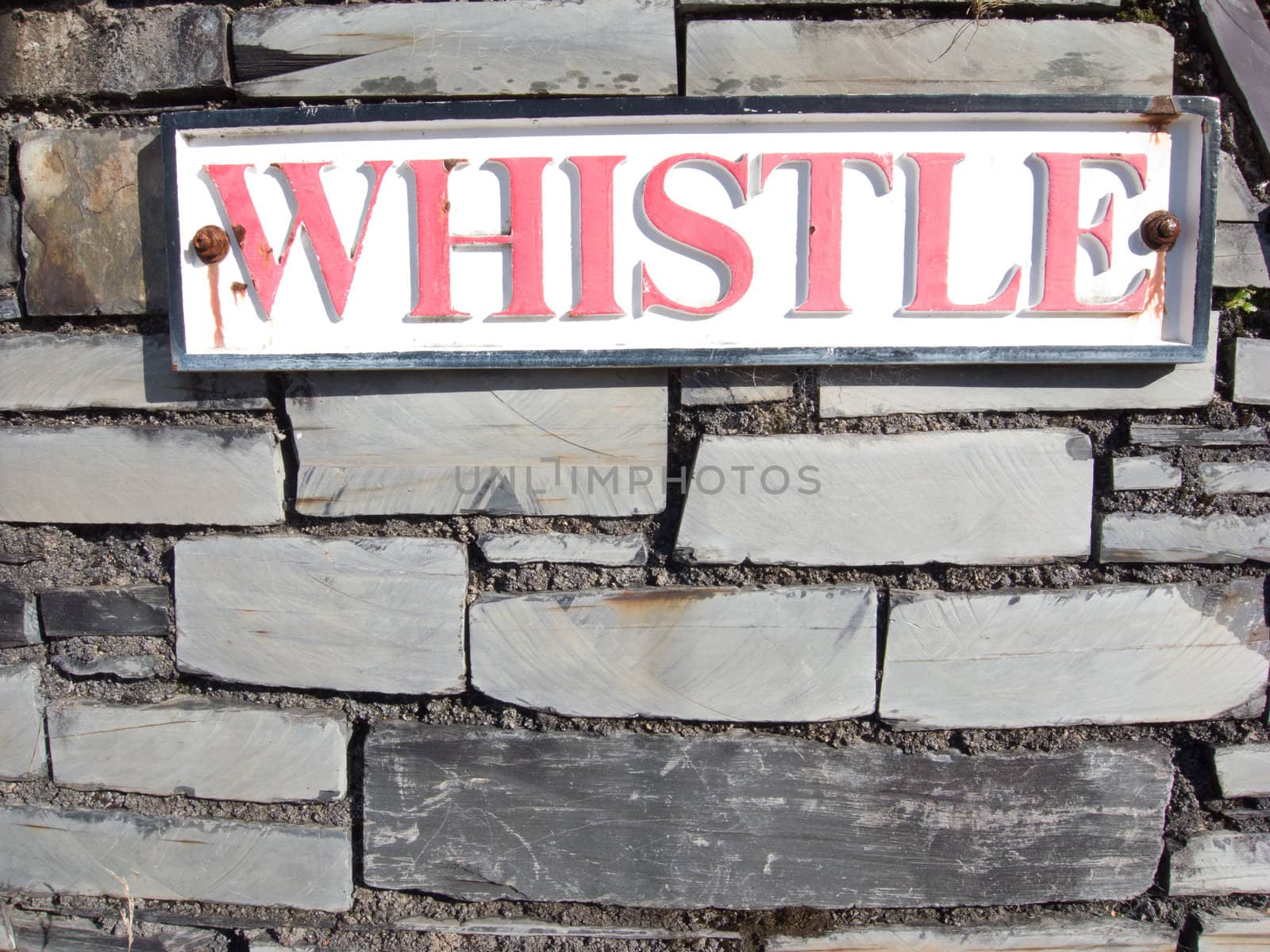 A sign with the word 'Whistle' in red paint on a white background with black border on a slate stone wall.