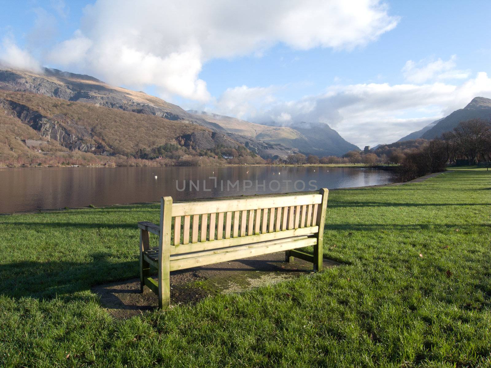 A wooden bench on green grass next to lake Padarn with a view of mountains in the Snowdonia national park, Wales, UK.