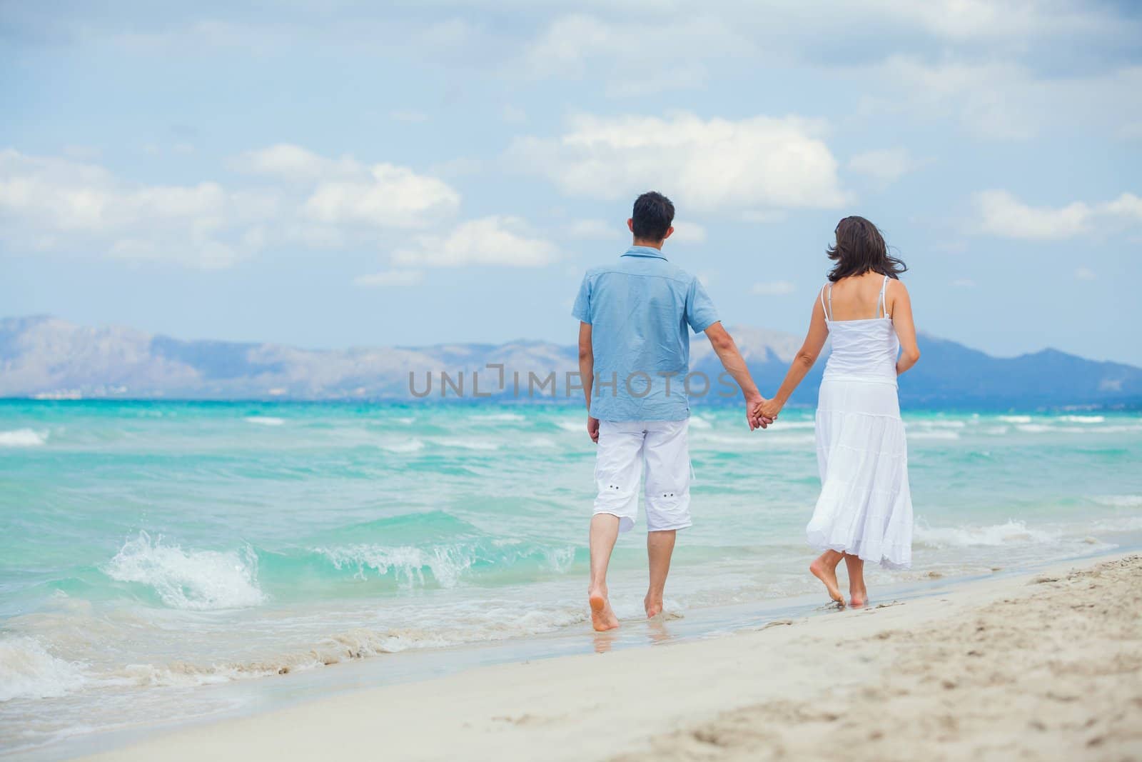 Back view of happy young couple walking on a deserted tropical beach with bright clear blue sky