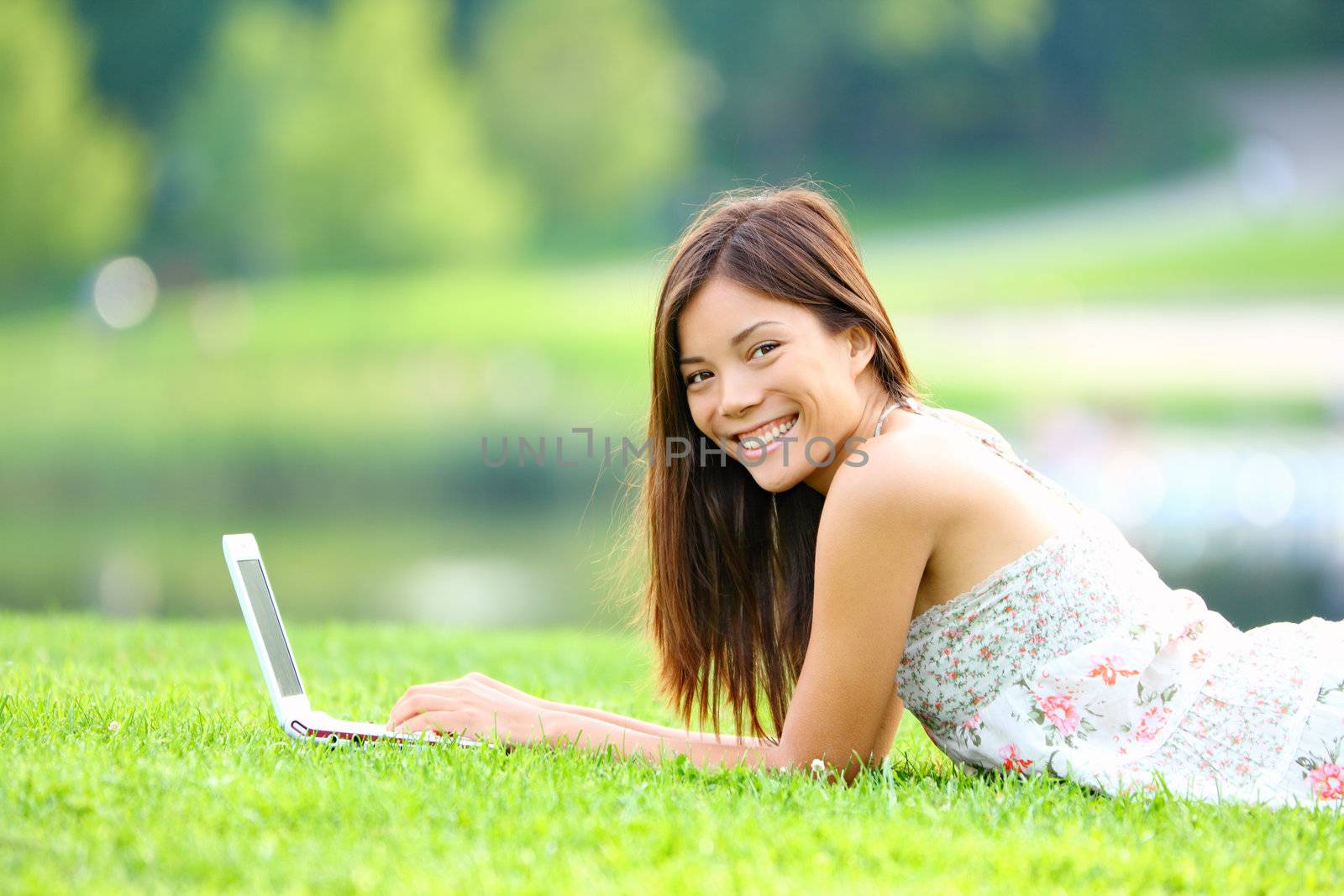 Girl in park on laptop. College student studying on laptop in summer or spring city park. Beautiful happy mixed race Caucasian / Asian chinese university student lying in the grass outside.
