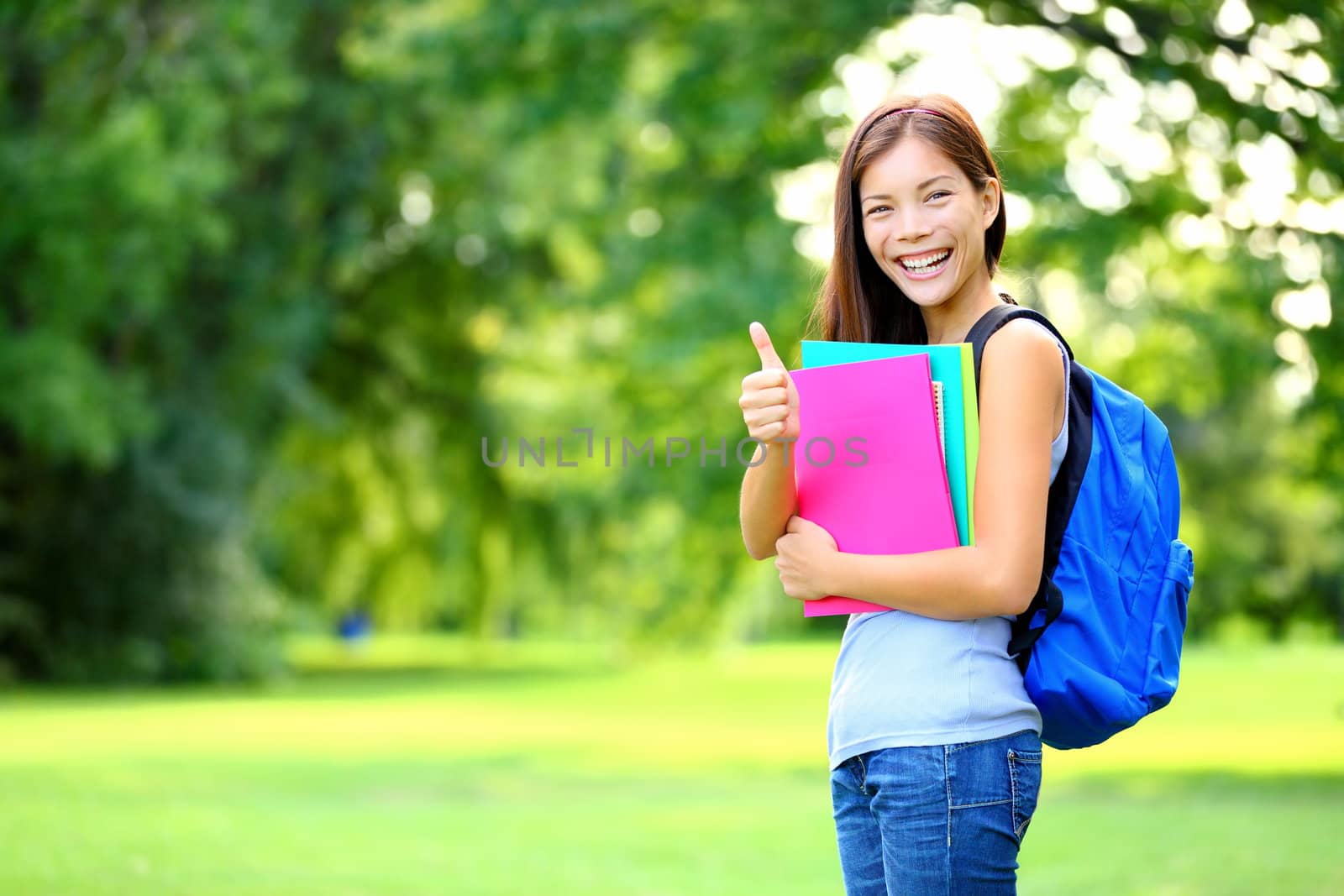 Student in park showing happy thumbs up success sign. Mixed race Asian Caucasian woman university student holding books outside.