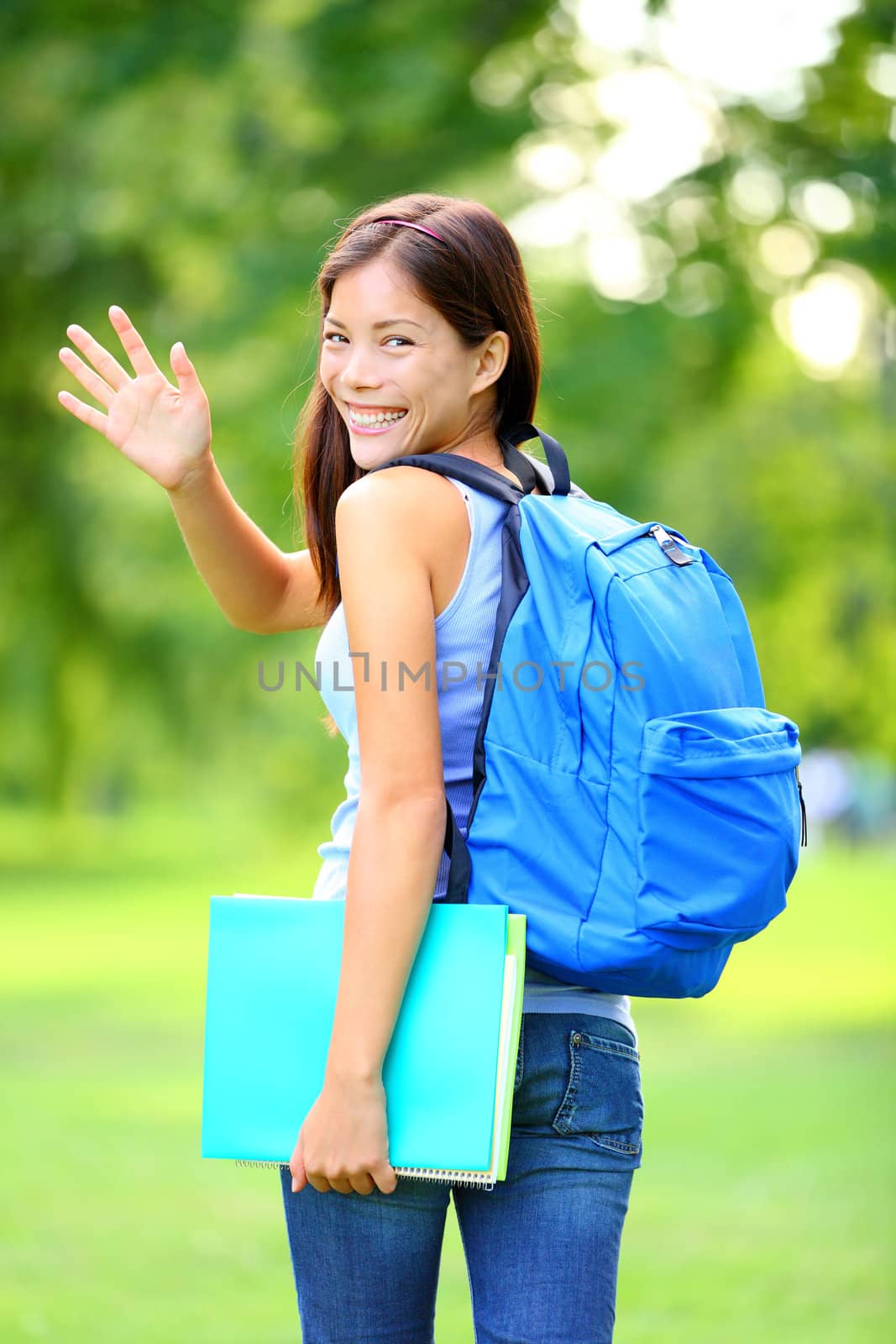 Woman student waving hello walking with school back in park smiling happy. Young female college or university student of mixed Asian / Caucasian race outside.