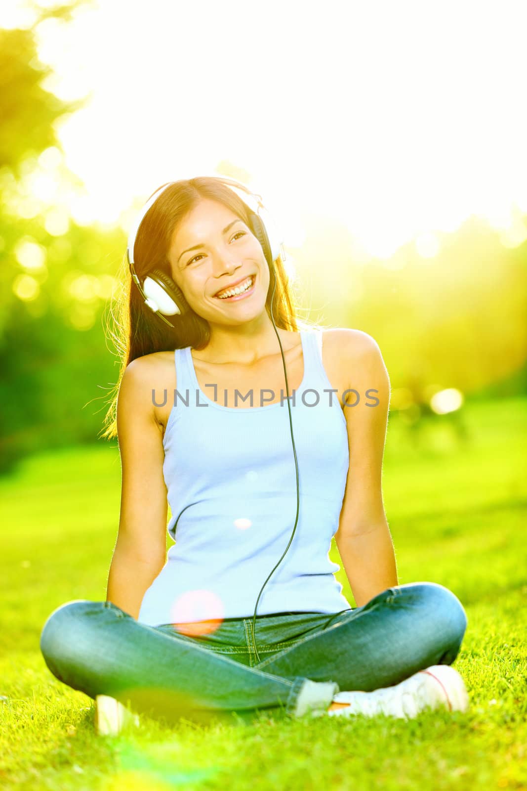 Woman listening to music on headphones in park smiling happy sitting in sunshine. Multiracial Caucasian / Chinese Asian girl.