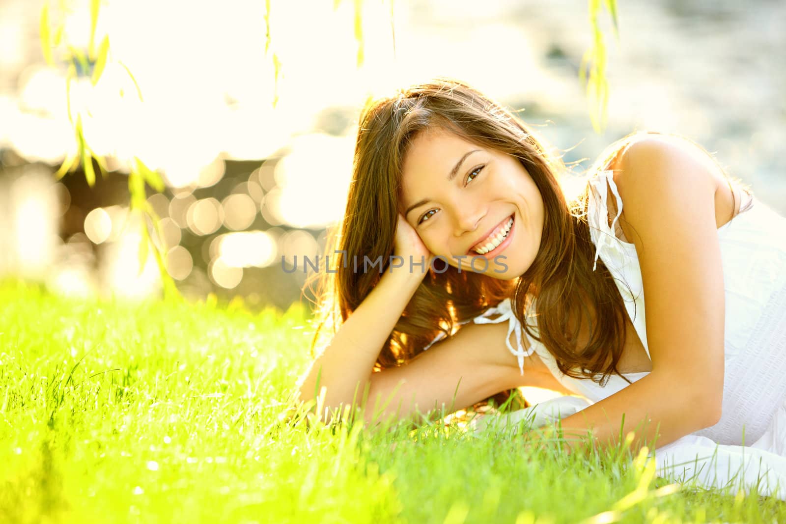 Summer girl in grass smiling happy. Lifestyle image of beautiful young woman in summer dress lying joyful in park on sunny sunshine day. Mixed race Caucasian / Asian Chinese female model.