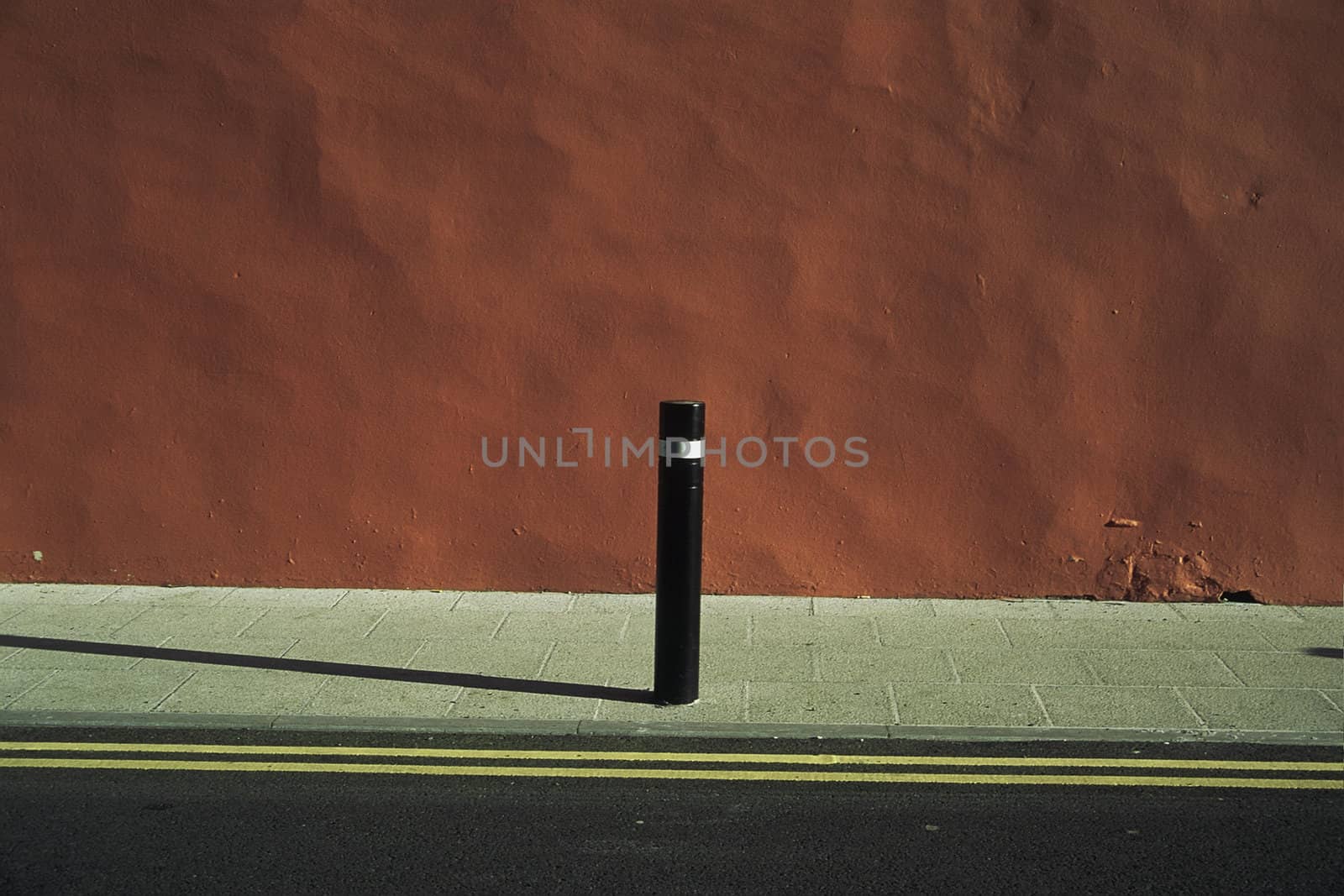 An urban abstract with road, pavement, yellow lines, orange dappled wall and a post with a shadow.