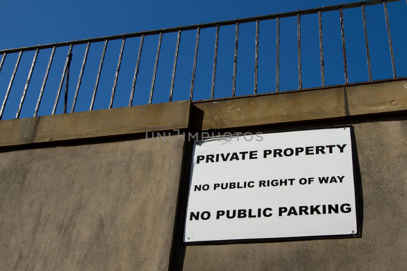 A white square sign with the message 'PRIVATE PROPERTY, NO PUBLIC RIGHT OF WAY and NO PUBLIC PARKING' on a wall with railings against a blue sky.