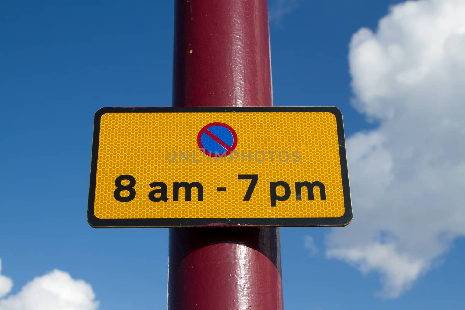 An oblong yellow sign with black border, the symbol for no parking and the time for no parking attached to a red post against a blue cloudy sky.