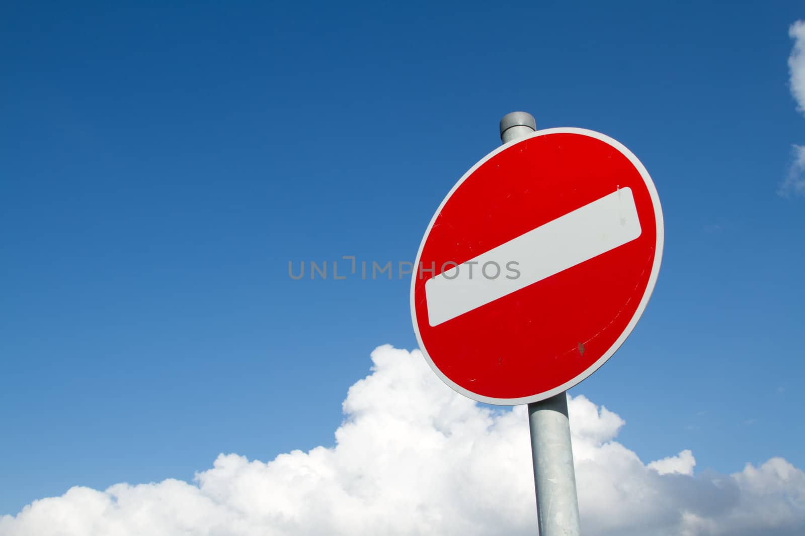 A circular red sign with a white bar indicating 'NO ENTRY' on a grey metal post against a blue cloudy sky.