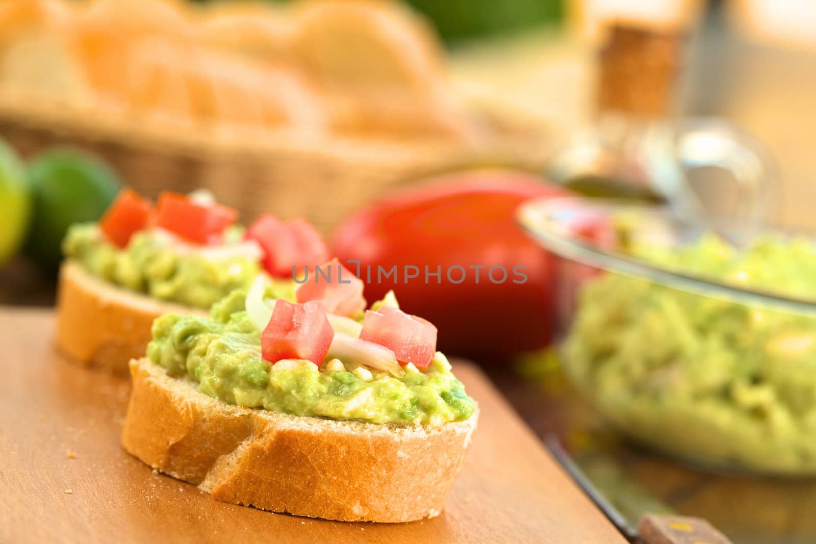 Snack of baguette slices with avocado cream, tomato and onion on wooden cutting board with ingredients in the back (Selective Focus, Focus on the front of the avocado cream on the first baguette slice)