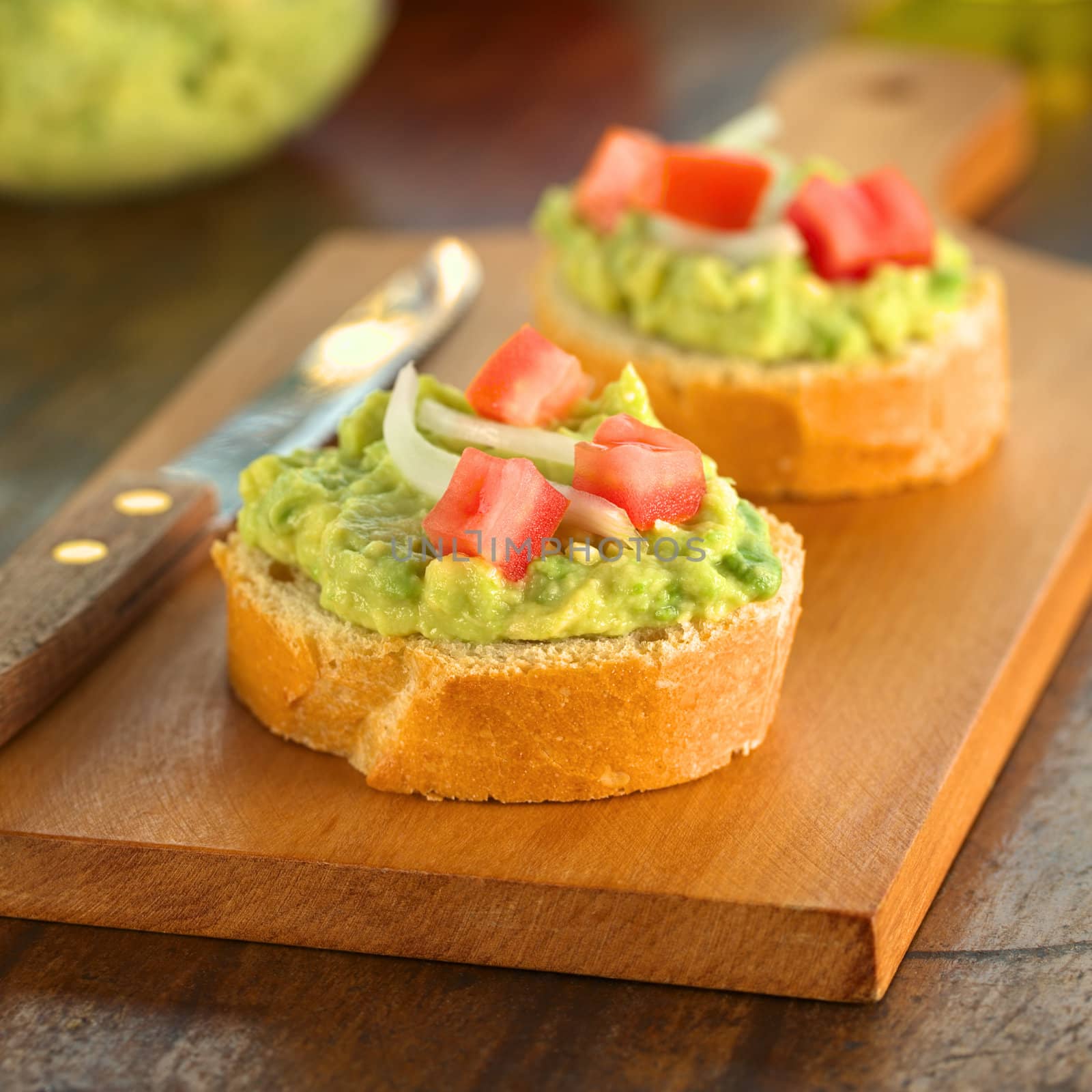 Snack of baguette slices with avocado cream, tomato and onion on wooden cutting board (Selective Focus, Focus on the front of the avocado cream on the first baguette slice)