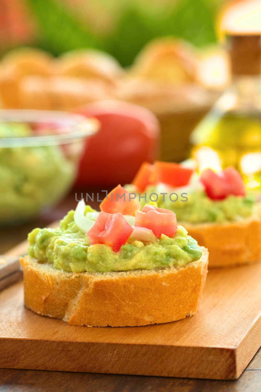Snack of baguette slices with avocado cream, tomato and onion on wooden cutting board with ingredients in the back (Selective Focus, Focus on the front of the avocado cream on the first baguette slice)