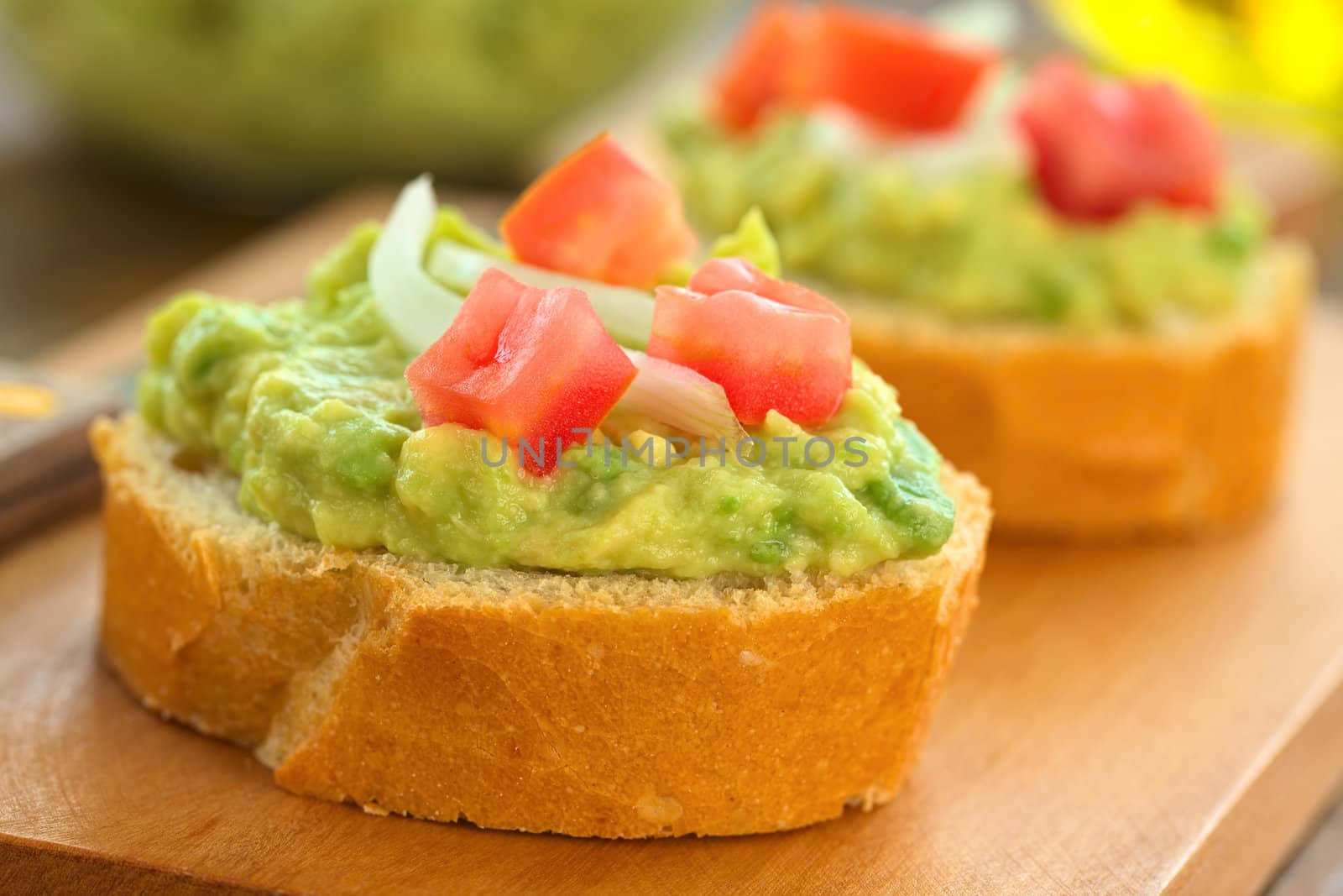 Snack of baguette slices with avocado cream, tomato and onion on wooden cutting board (Selective Focus, Focus on the front of the avocado cream on the first baguette slice)