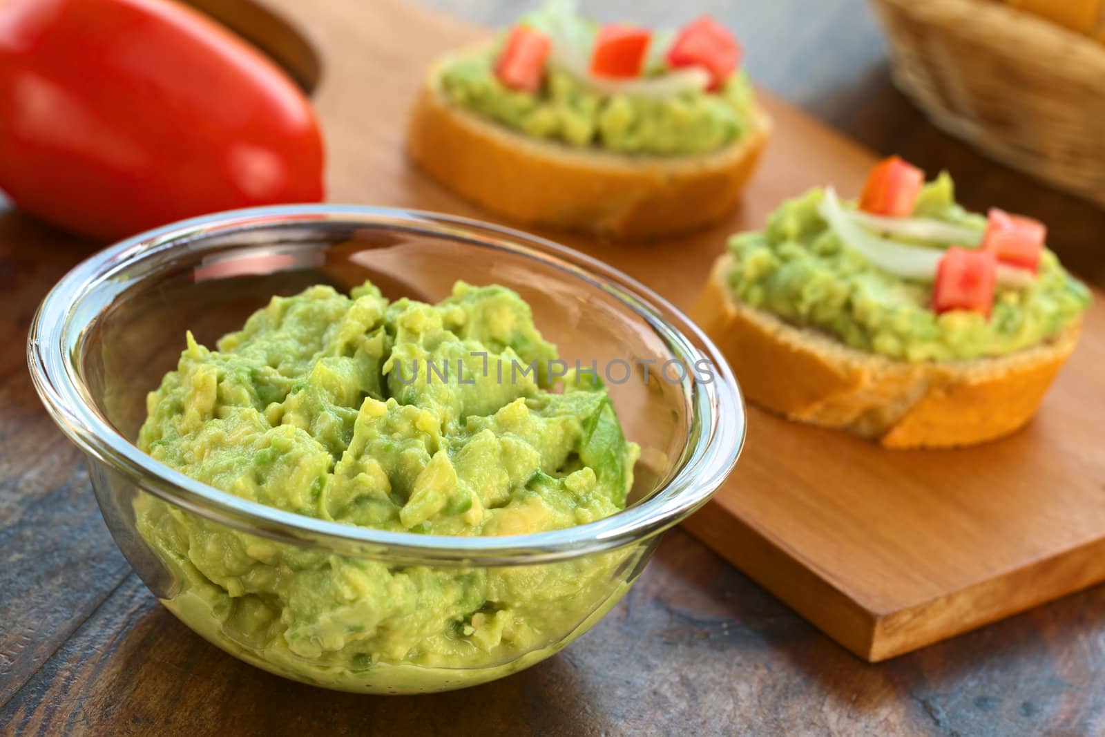 Fresh avocado cream in glass bowl with snacks of baguette with avocado, tomato and onion in the back (Selective Focus, Focus one third into the avocado cream)