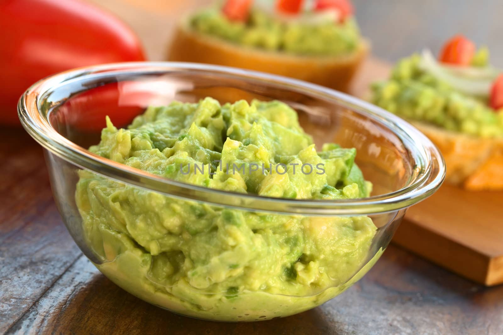 Fresh avocado cream in glass bowl with snacks of baguette with avocado, tomato and onion in the back (Selective Focus, Focus one third into the avocadocream)