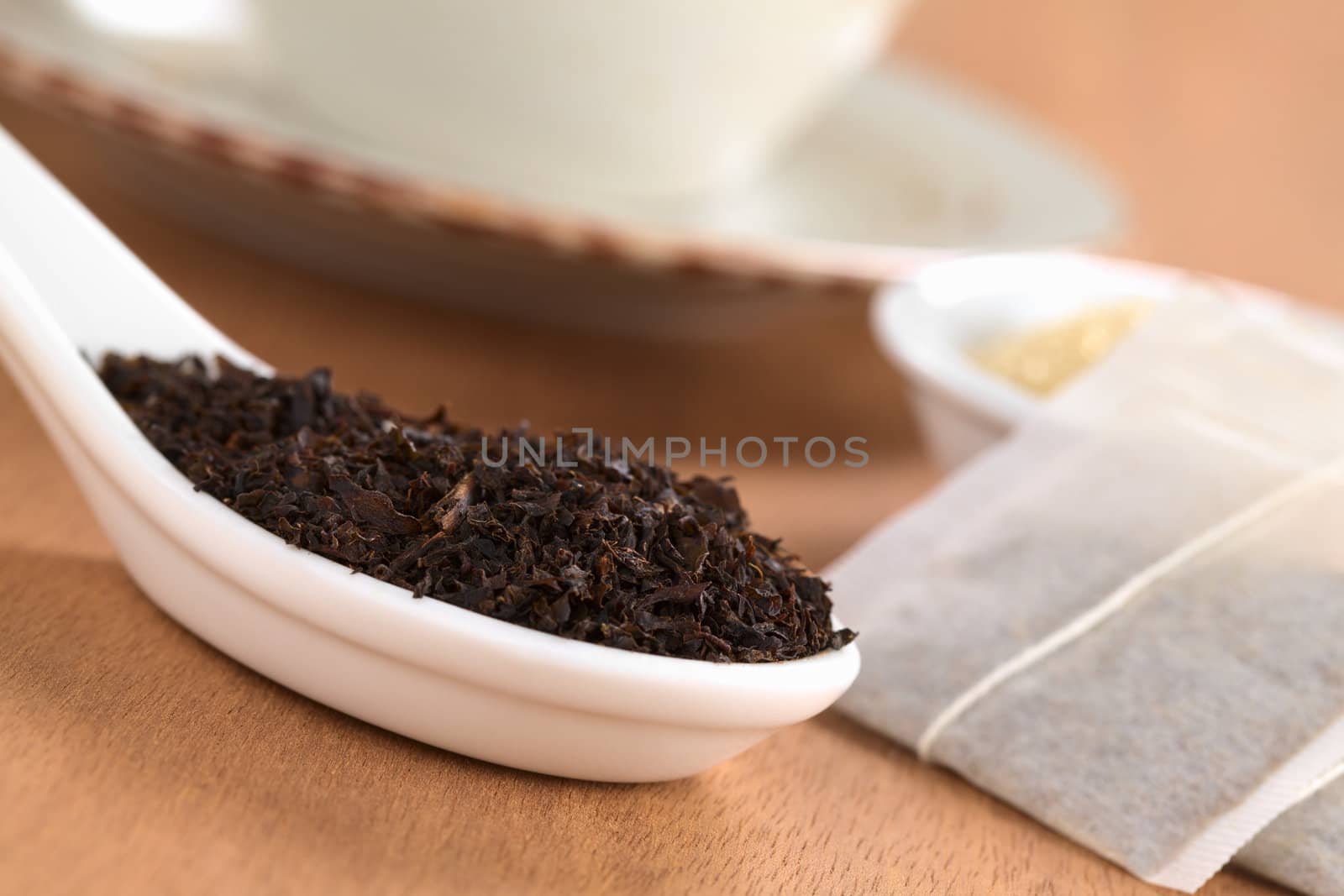 Dried loose black tea on ceramic spoon with tea bags, brown sugar and a cup in the back photographed on wood (Selective Focus, Focus one third into the leaves on the spoon)