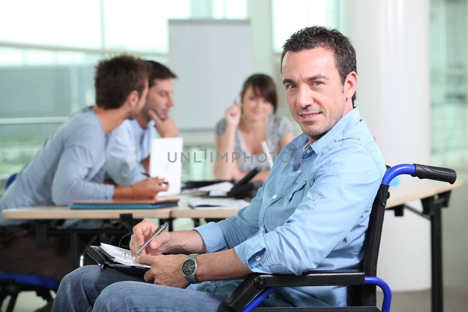 Office worker in a wheelchair with colleagues in the background by phovoir