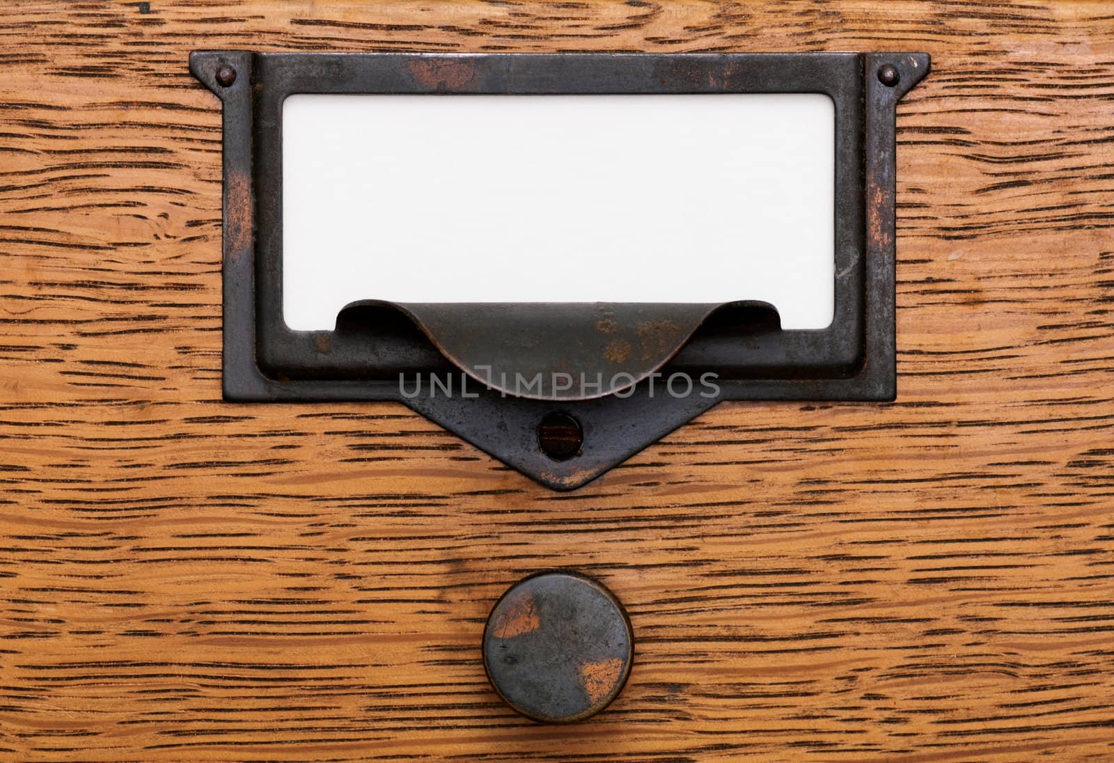 Close up of a blank, white label in a grungy, tarnished brass label holder and drawer pull on an oak filing cabinet drawer.