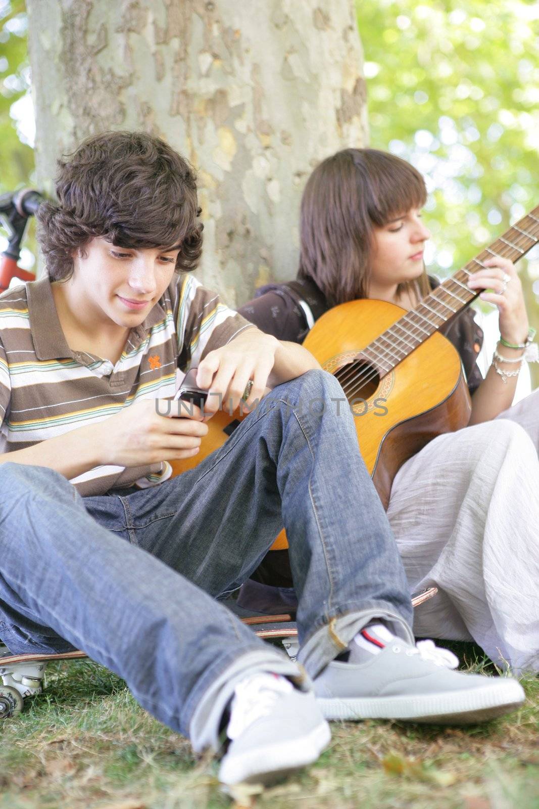 Two teens with guitar sat by tree