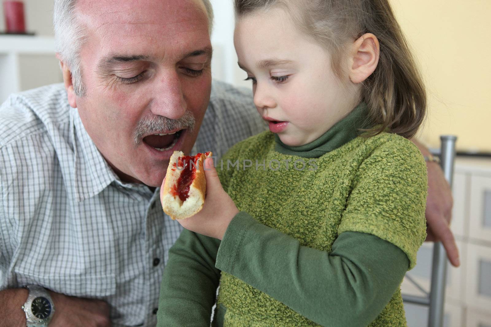 Granddaughter giving her grandfather a piece of bread with jam to eat by phovoir