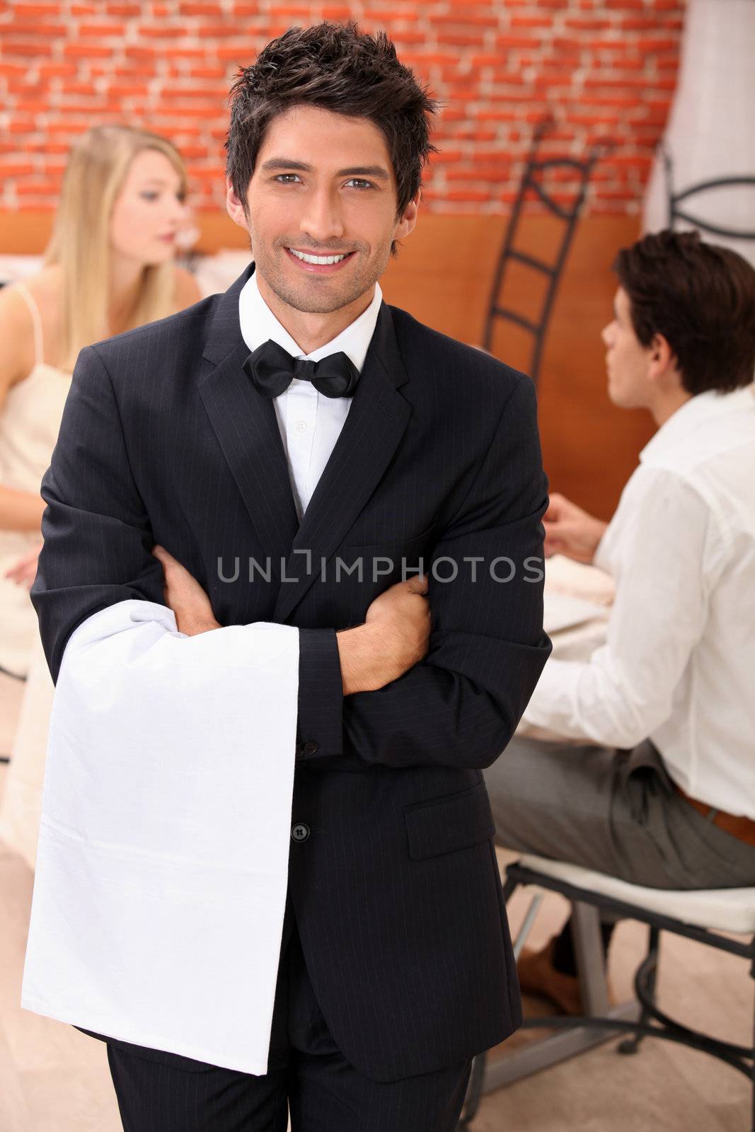 well dressed waiter facing the camera, behind a couple is dining at restaurant