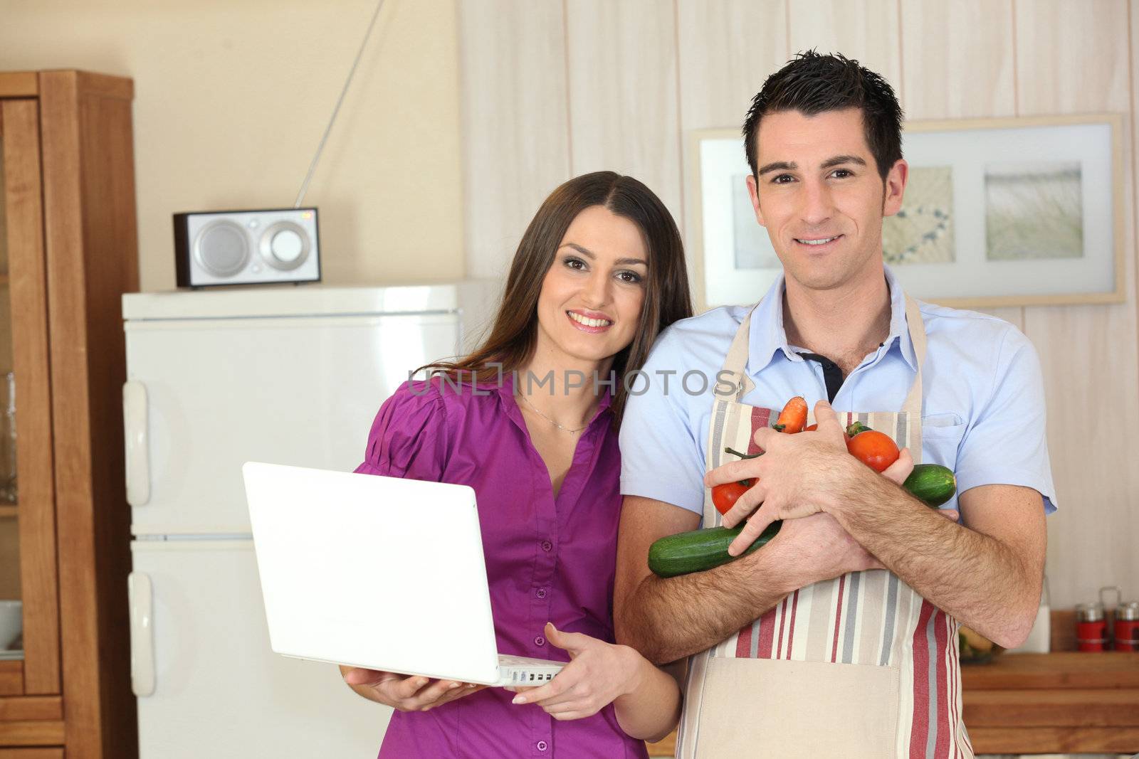 Couple in a kitchen with a laptop computer and arms full of vegetables by phovoir