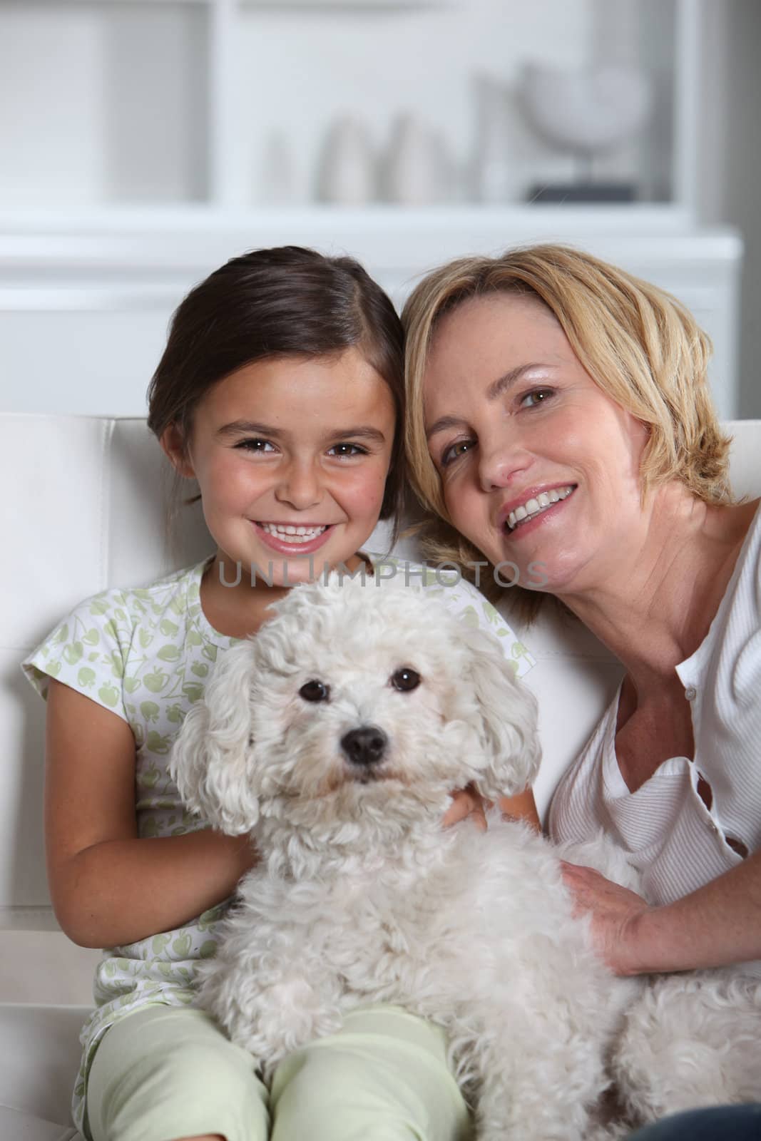 Mother, daughter and white dog sitting in a white living room by phovoir