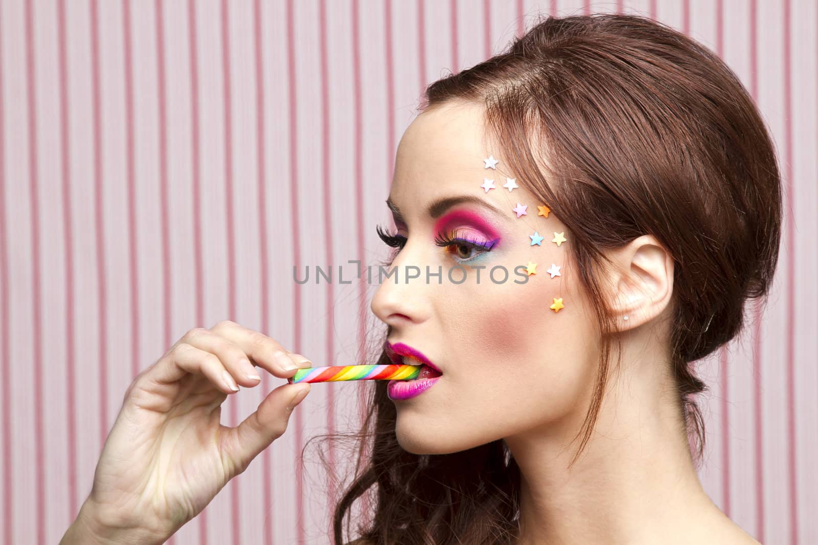 Young woman with colorful makeup and star candy glued to her face, bitting a candy cane