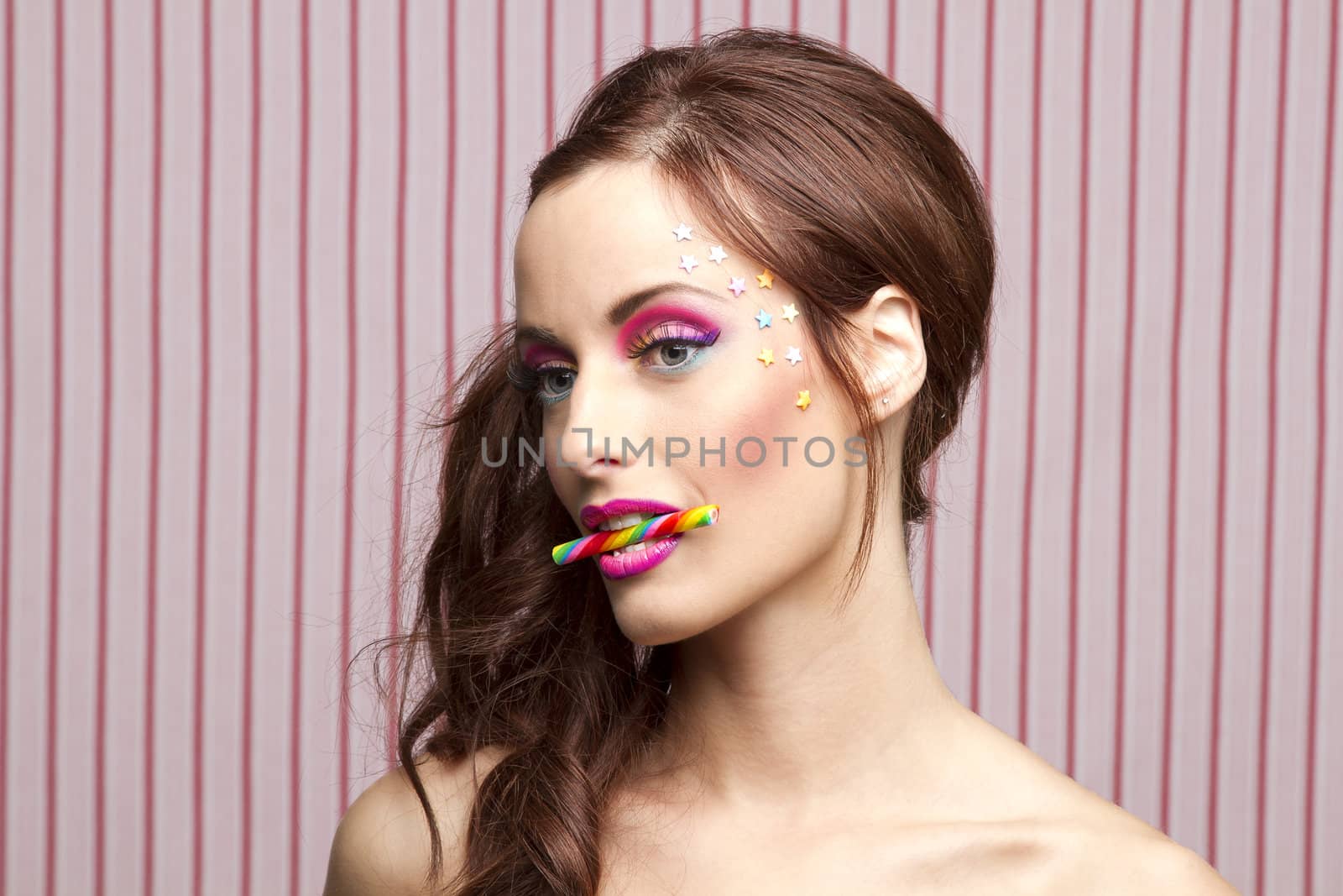 Young woman with colorful makeup and star candy glued to her face, bitting a candy cane