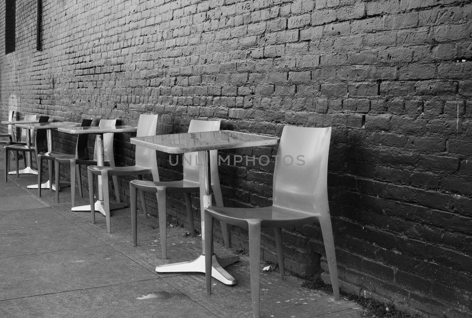 Black and white image of brushed aluminum tables and chairs on a sidwalk against a black brick wall